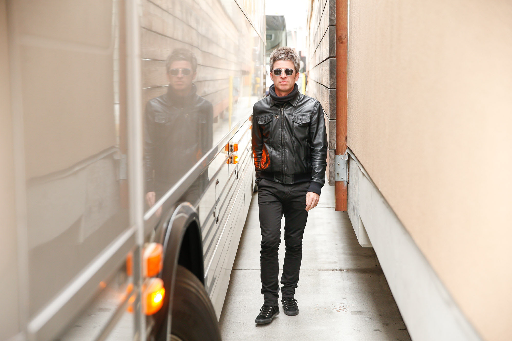 Noel Gallagher walks in between a bus and a building, wearing sunglasses and a leather jacket.