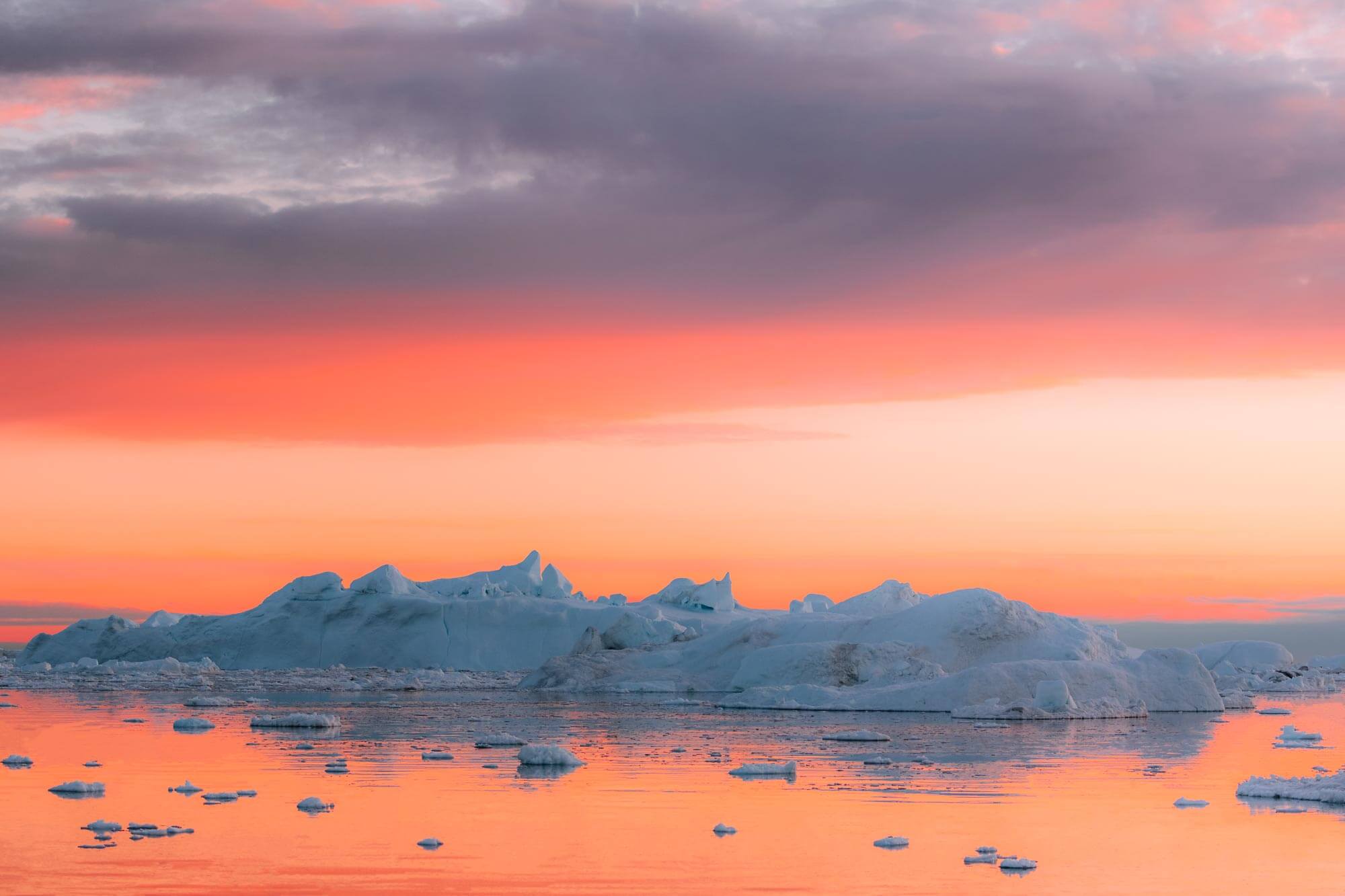 The ocean reflects an orange-red sunset and a range of icebergs in the background