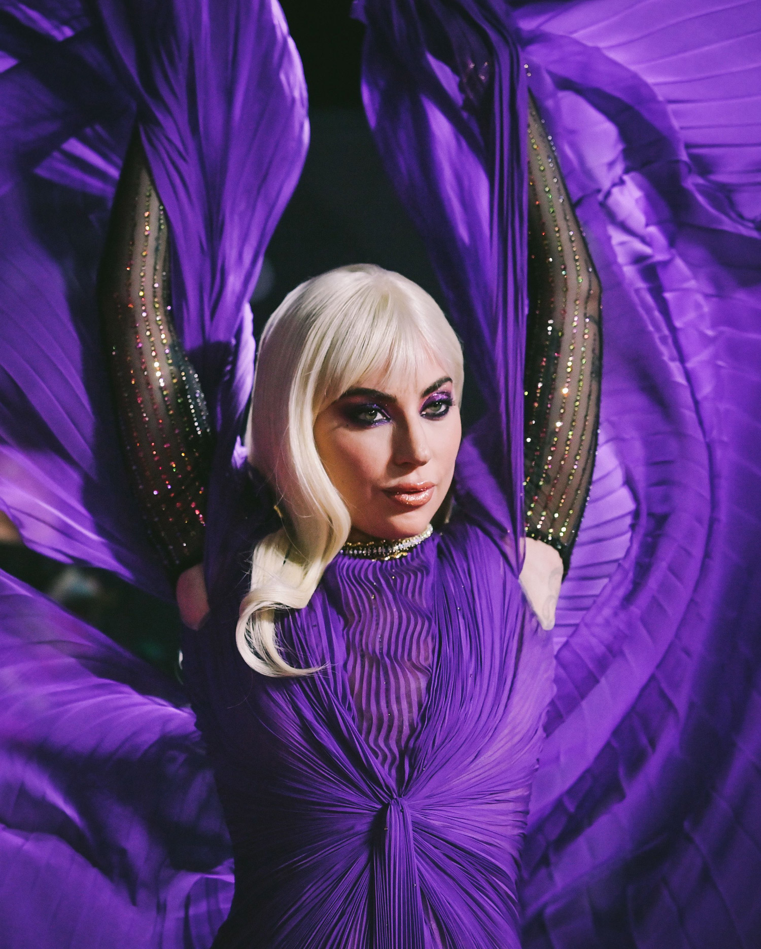 Lady Gaga wearing a purple dress stands with two arms in the air, with a long black glove on each arm.