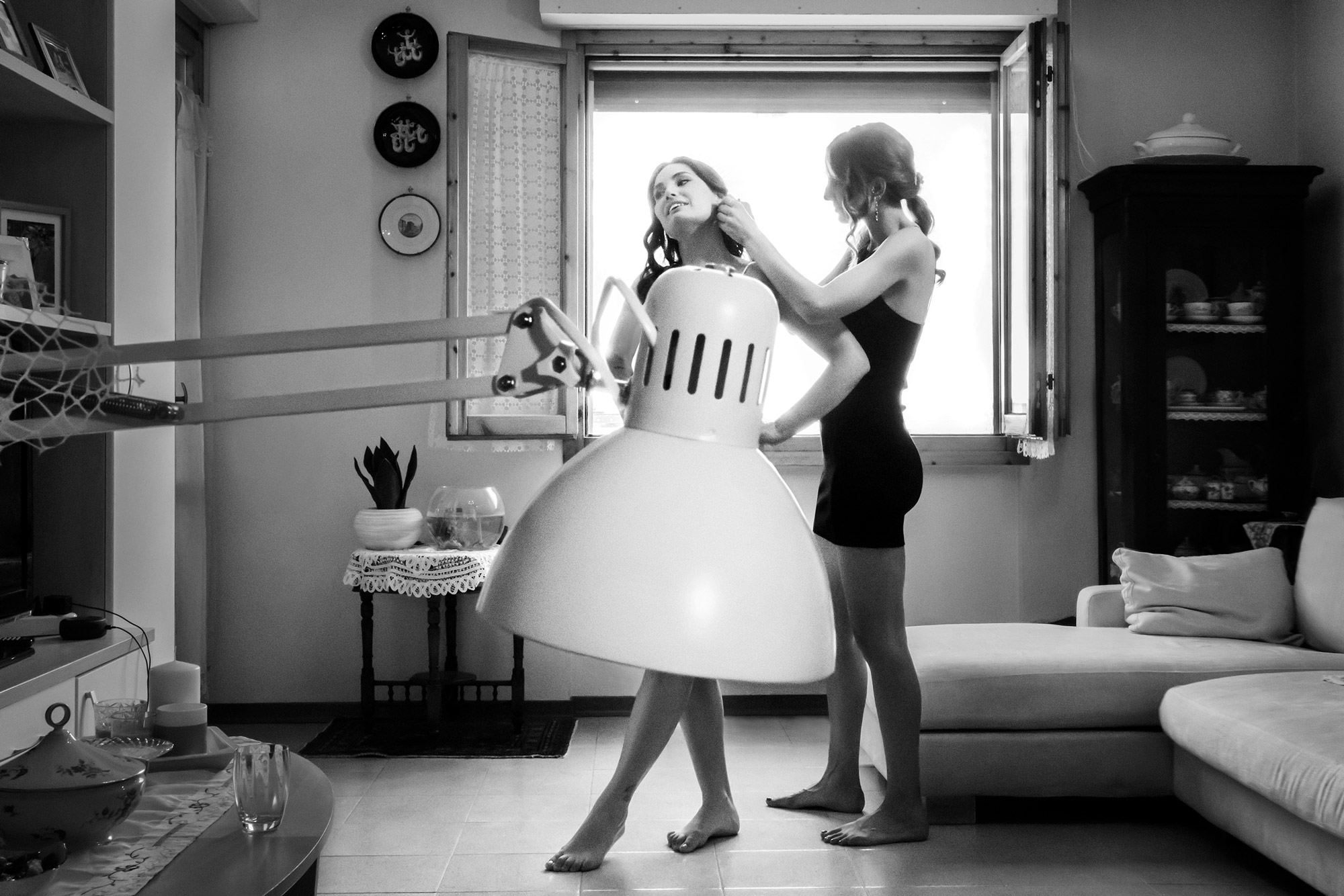 A black and white image of two women in a room in front of an open window, surrounded by a sofa and cupboards. A table lamp covers one of the women, as the other one fixes her earring.