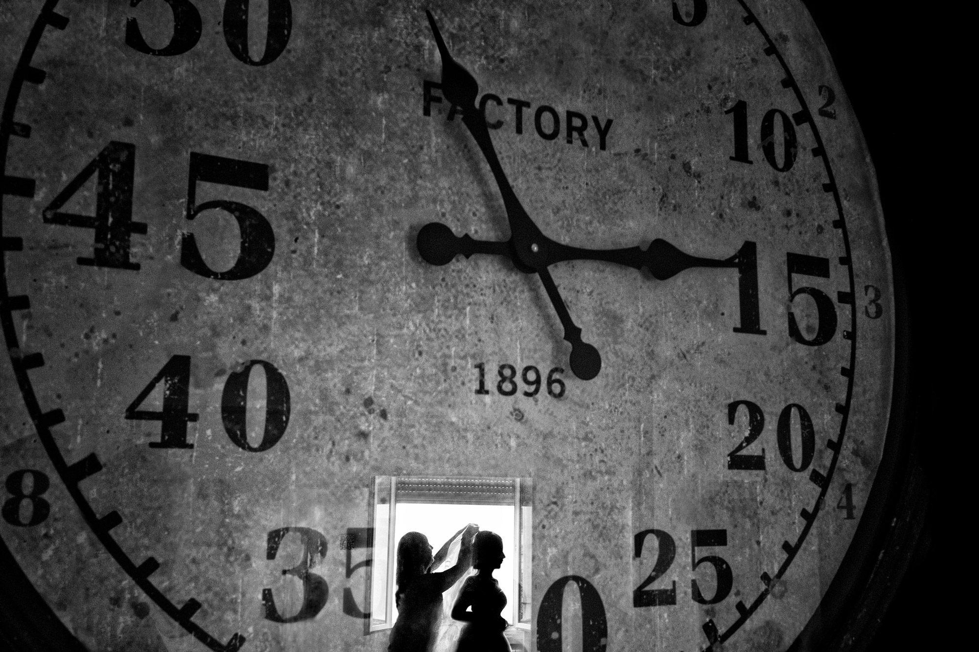 A black and white image of a giant clock, at the bottom of the clock there is an open window with two women in silhouette, the woman on the left is attaching a veil to the other’s hair.