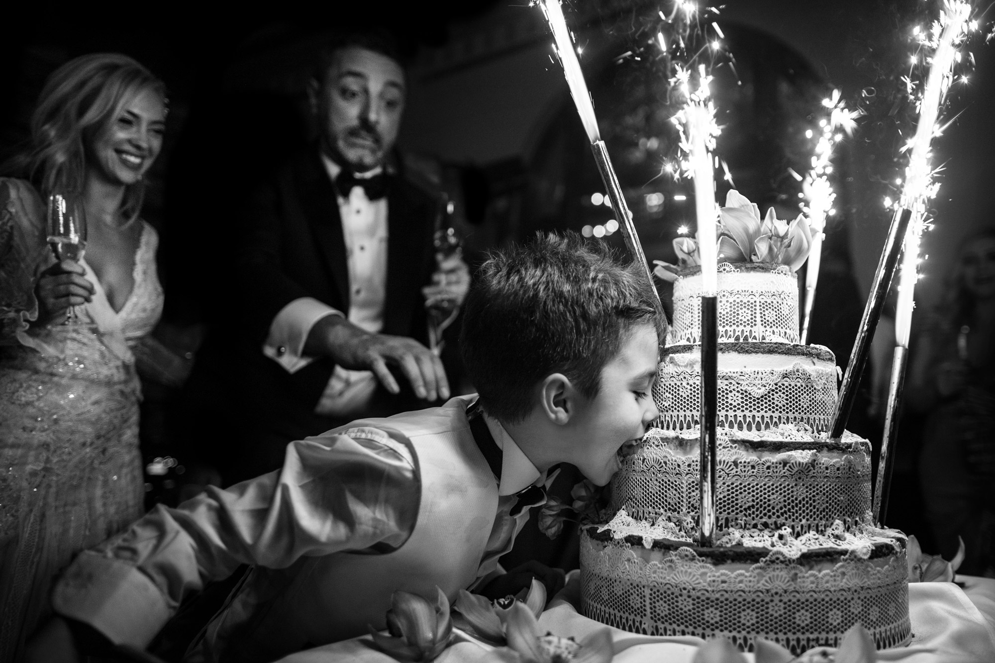 A black and white image of a boy leaning in to take a bit from a wedding cake that has sparklers on it. A bride and groom are in the background.
