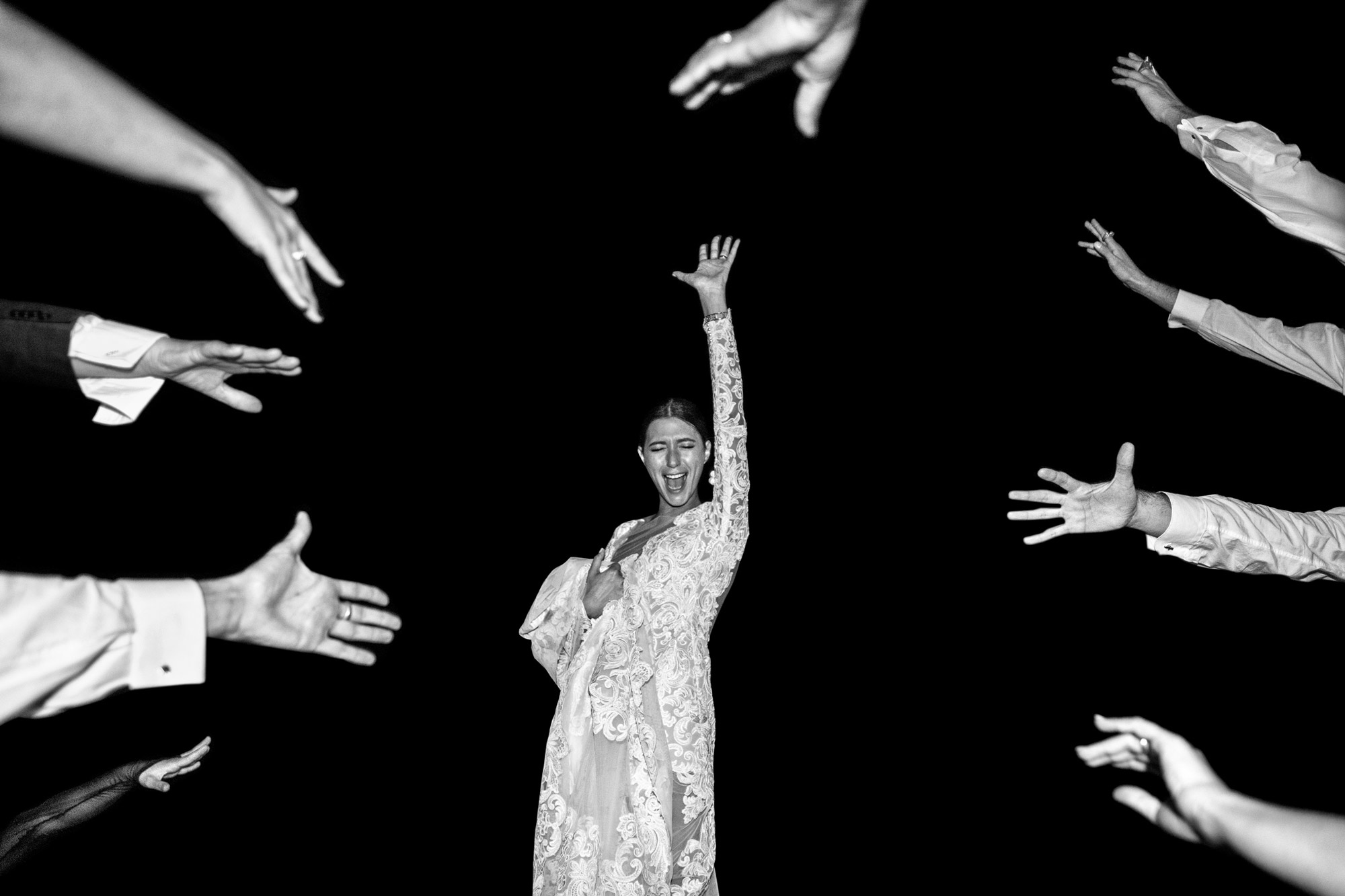 A black and white image of a bride wearing a white dress standing with one arm in the air, she is surrounded by a circle of arms.