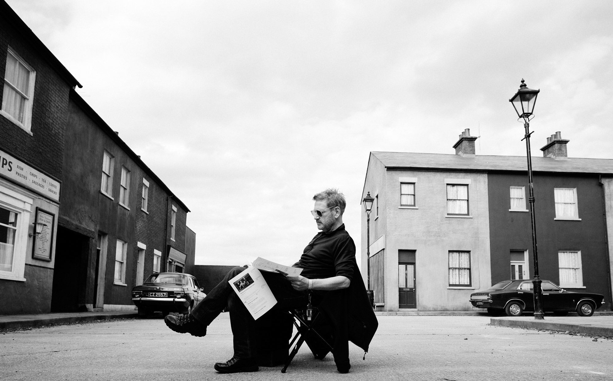 Kenneth Branagh reads a script, sitting on a chair on a film set.