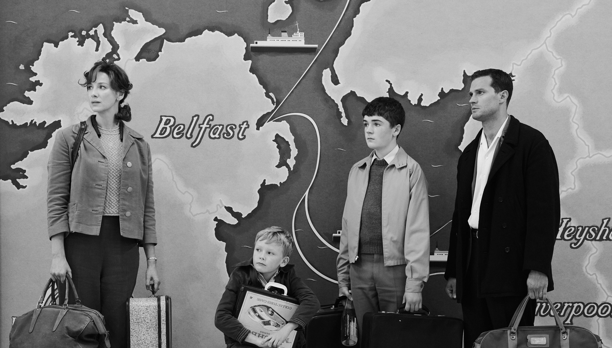 Four people carrying luggage stand in front of a large map of Britain.