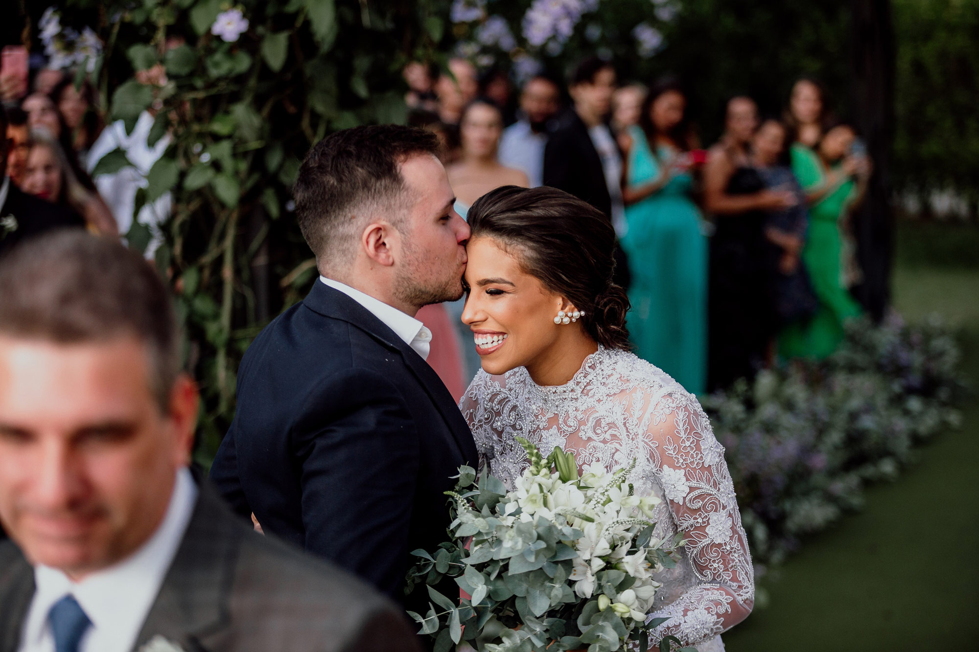 Groom wearing a suit kisses a smiling bride on the forehead