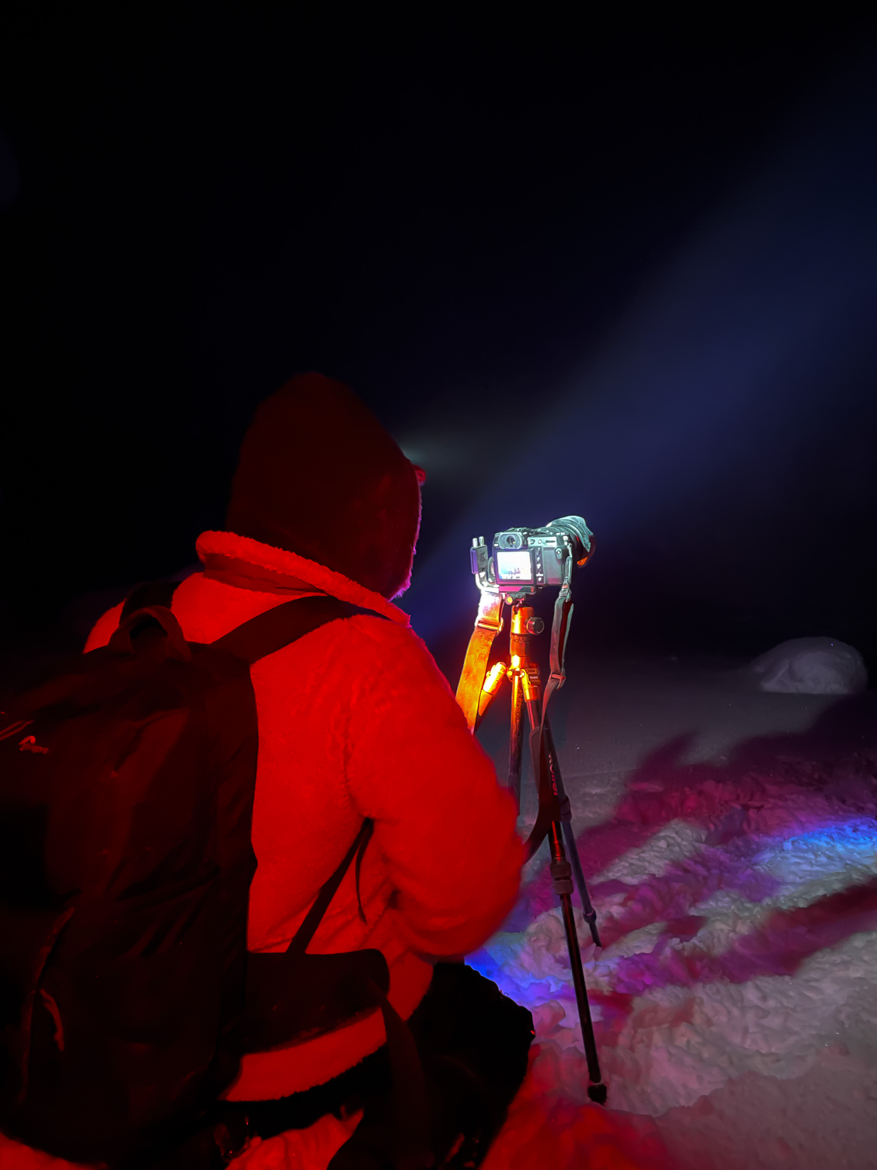 Foto einer Person mit roter Jacke, die ihre Kamera auf einem Stativ im Schnee betrachtet.