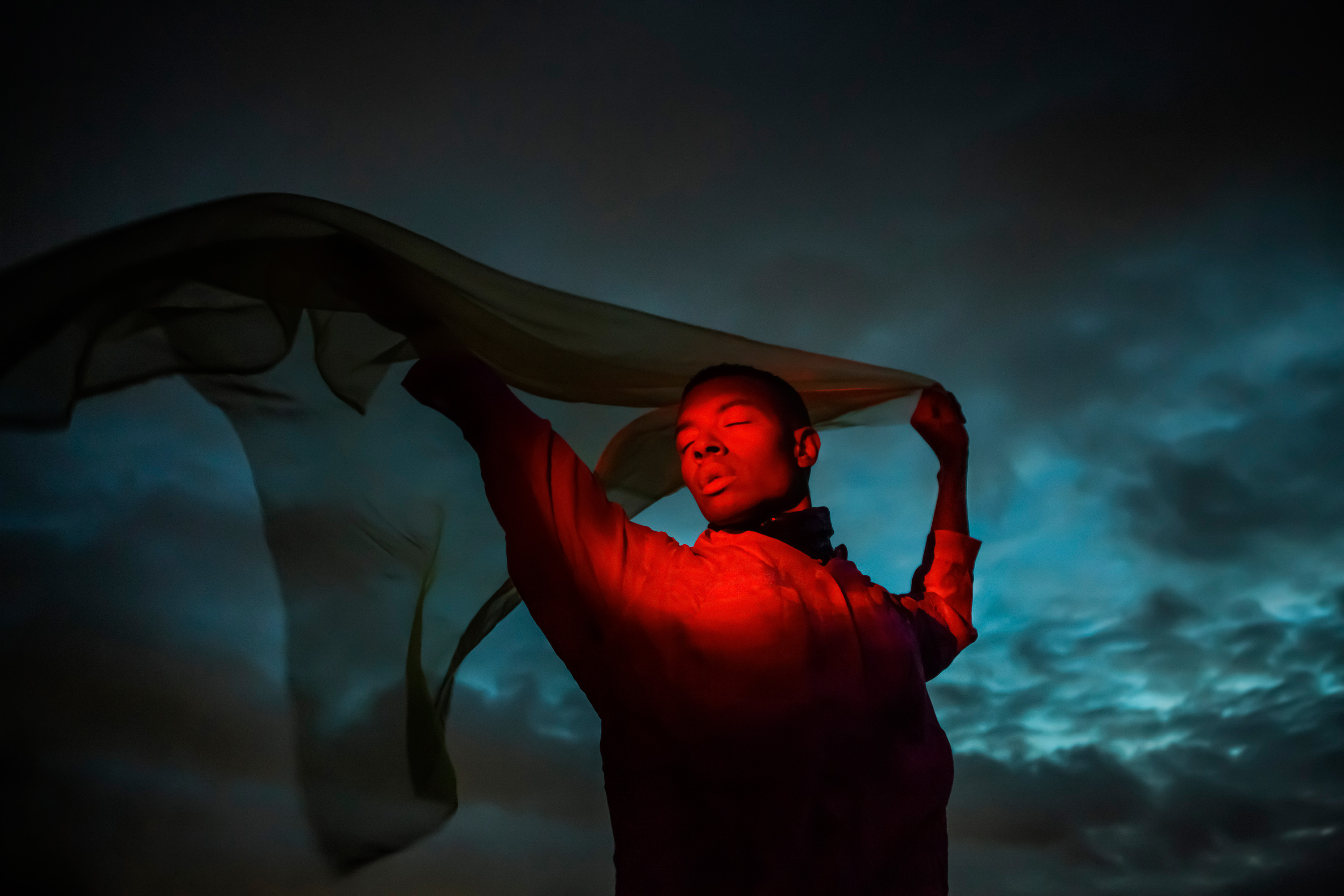 a cinematic shot of a man lit by artificial red light holding a see through fabric in the air in front of a dramatic blue sky