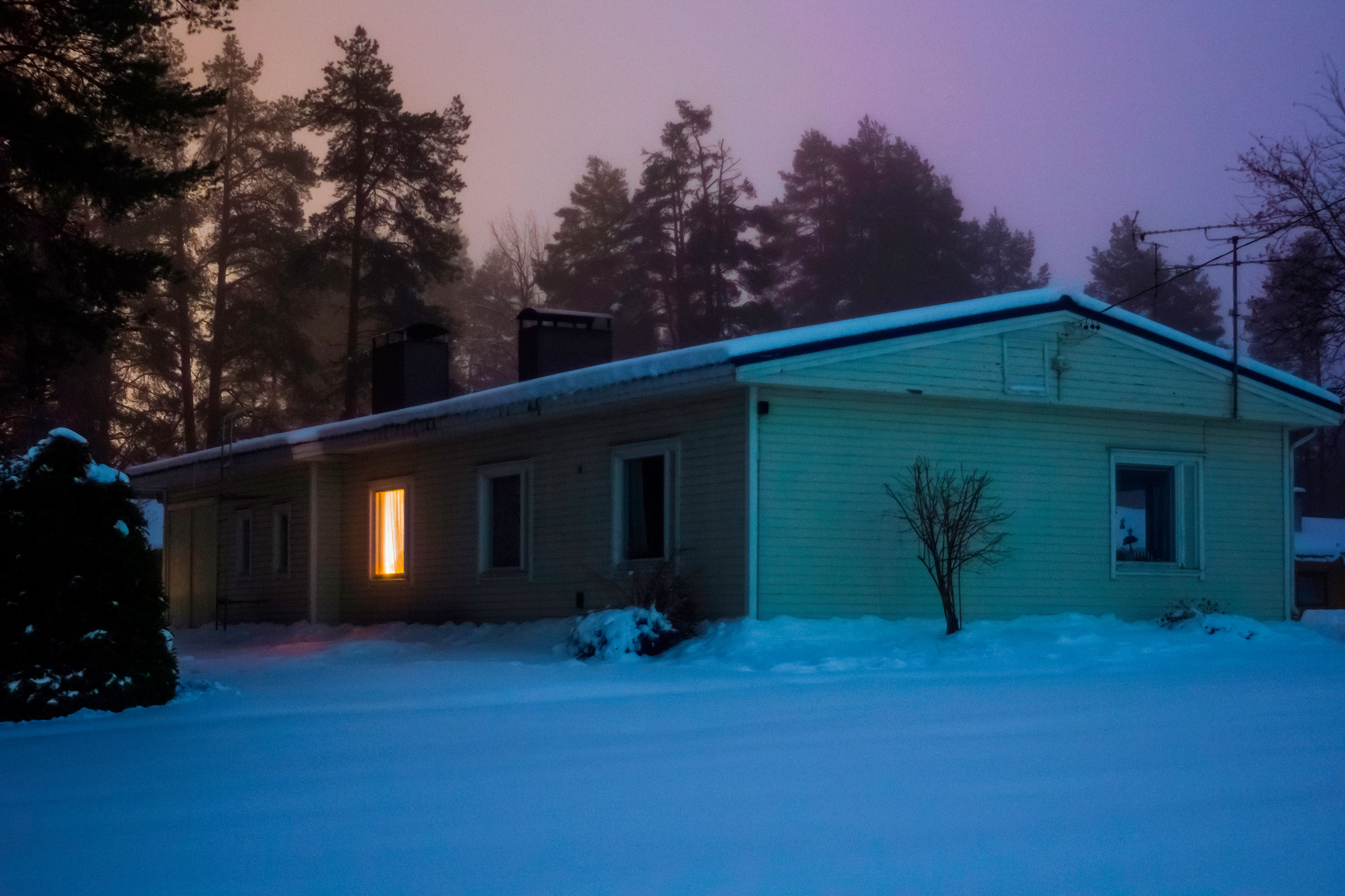 A house with a single tungsten light on, stands alone in a wintry landscape
