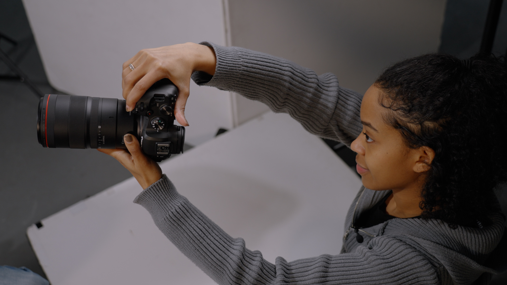 A photographer wearing a grey sweater holding a DSLR in a studio setting