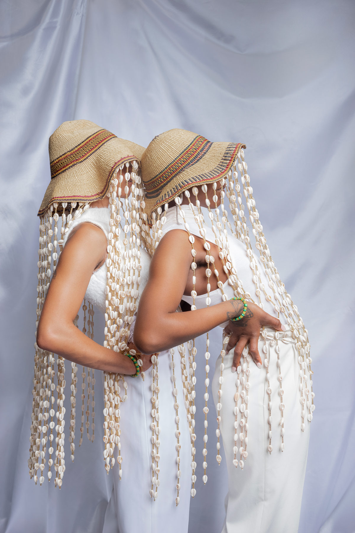 A pair of women wearing hats with strings of seashells attached hanging down to their stomachs being held by their hands