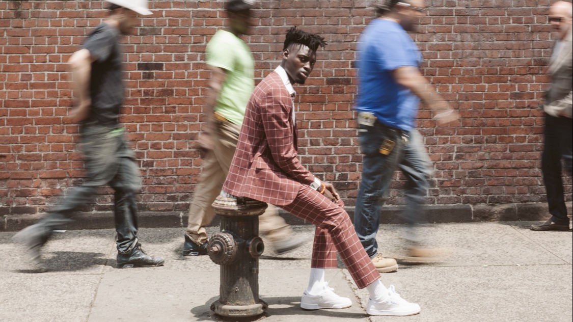 A black man wearing a dark red chequered suit and white trainers sits on a fire hydrant with a brick wall and blurry people walking past