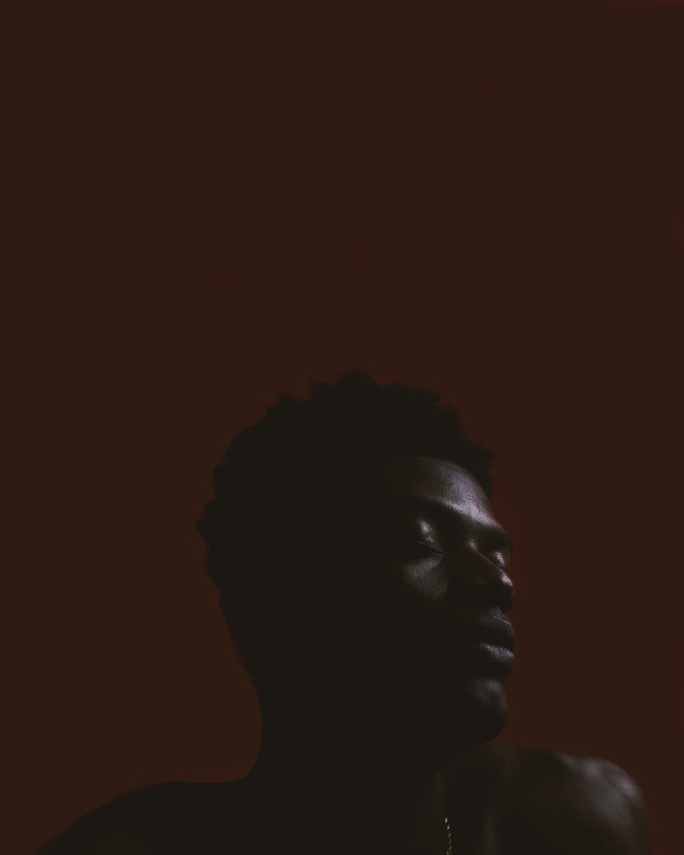 A black person with their eyes closed looks to the right of the camera against a dark brown background