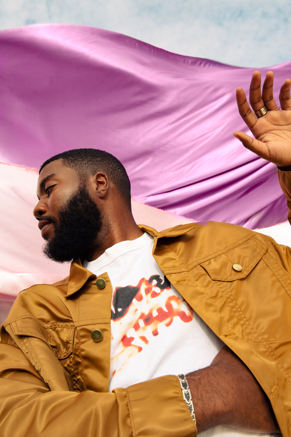A black man with a beard yellow jacket and gold ring holds their hand up while looking away from the camera a pink and white flag in the background
