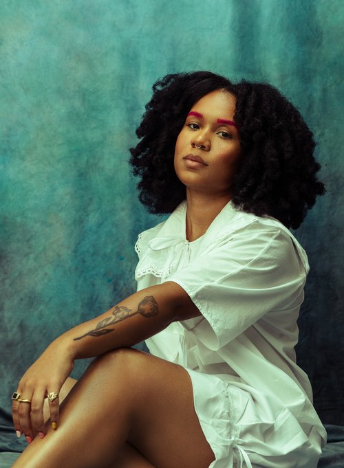 A Black woman with red eyebrows and a tattoo of a flower on their forearm wearing a white dress sitting in front of a turquoise background