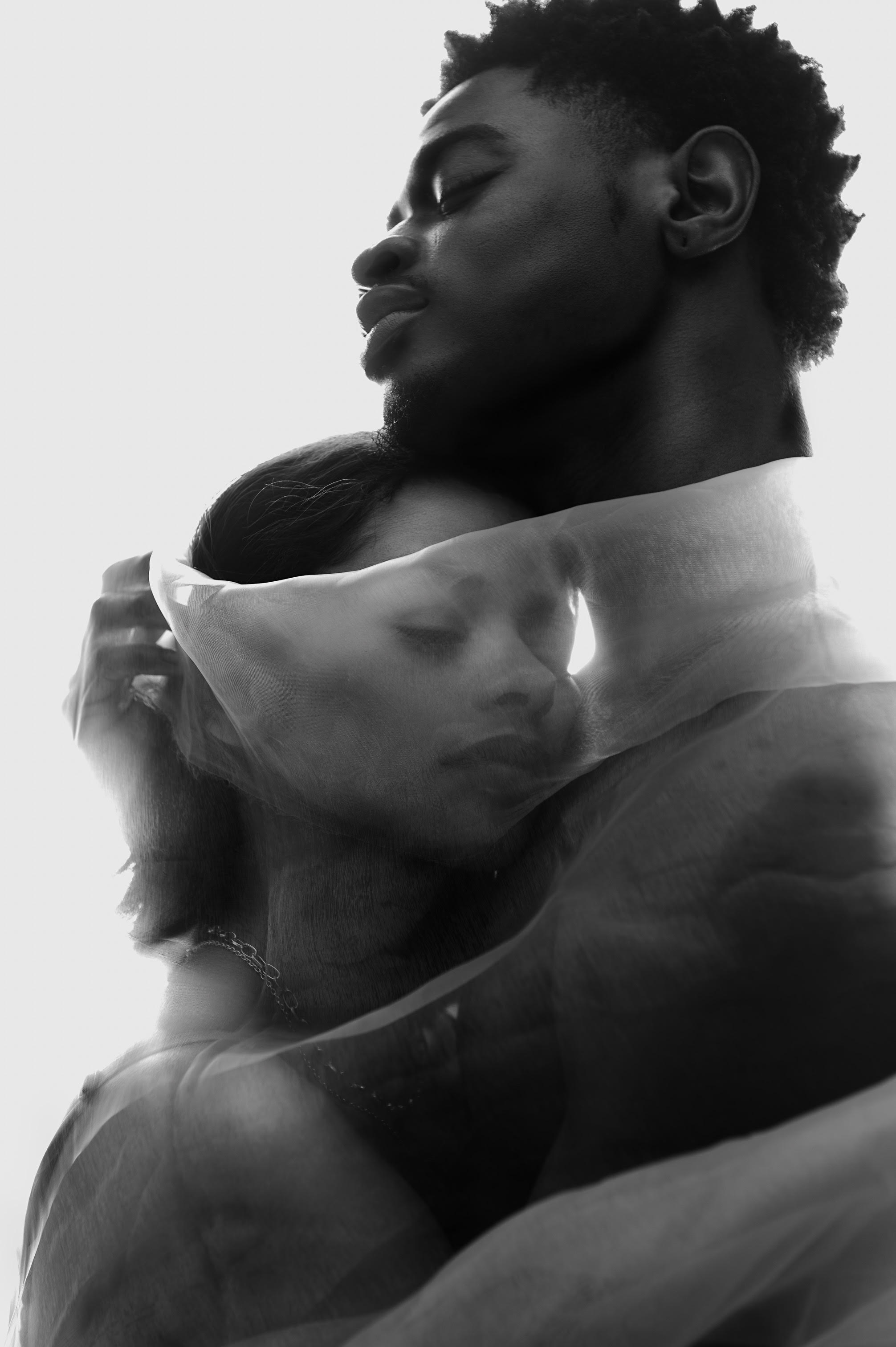 A black and white photo of a black man and woman embracing while wrapping themselves in semi transparent cloth