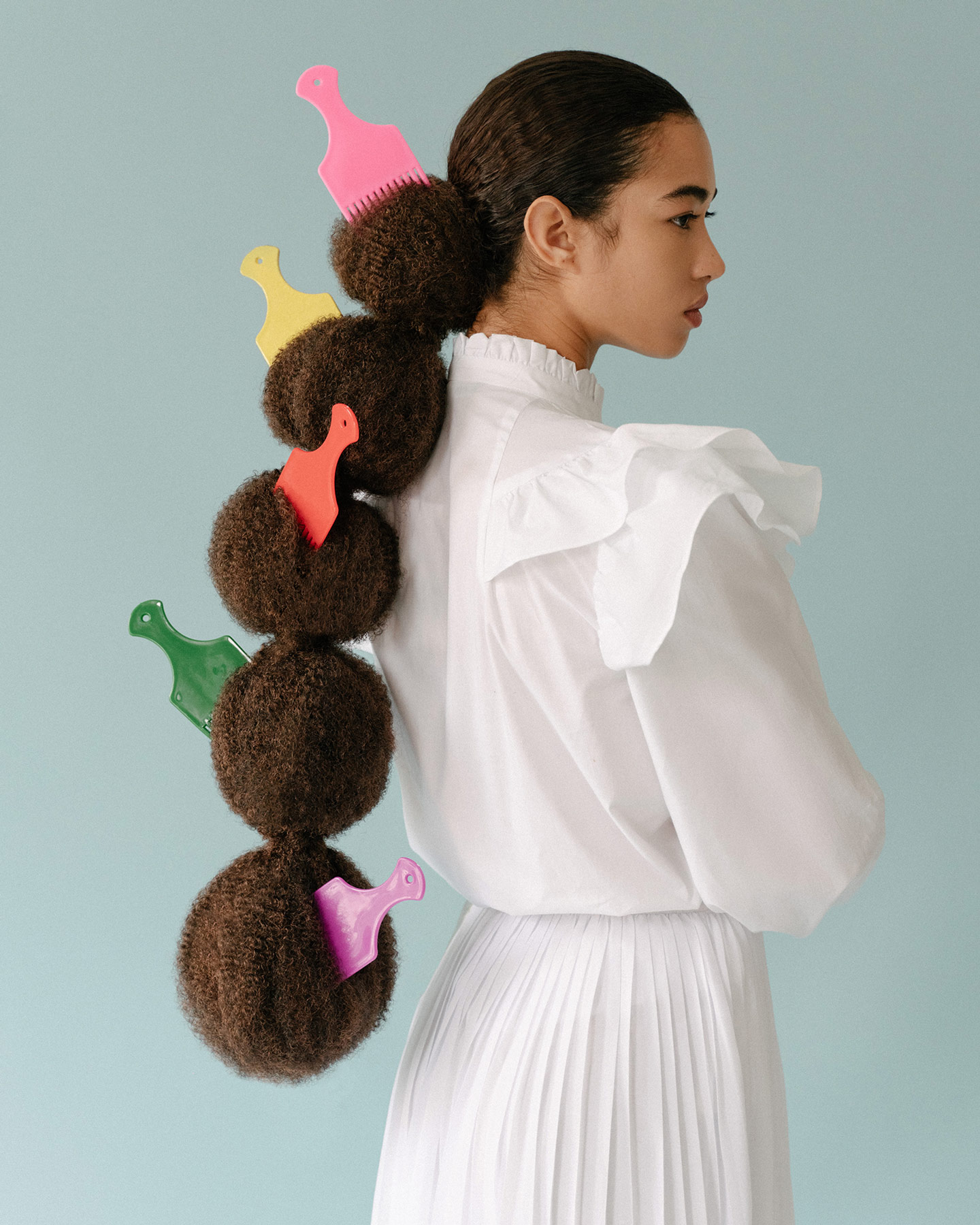 A black woman wearing a white dress with long hair shaped into five balls with an afro comb in each one standing with arms crossed against a light blue background