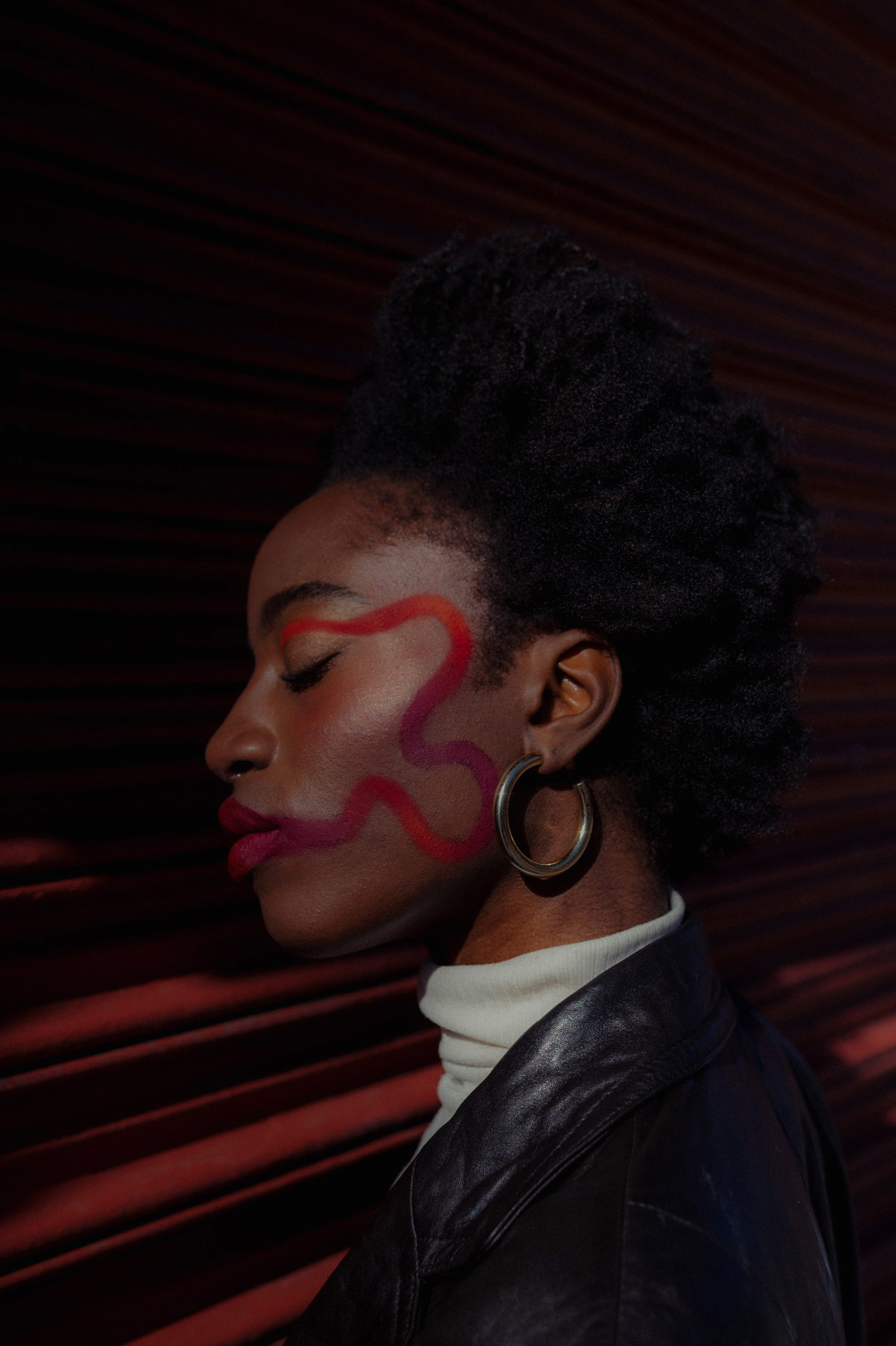 A Black woman wearing hooped earrings with red lipstick spiralling around her face looking to the left of the camera