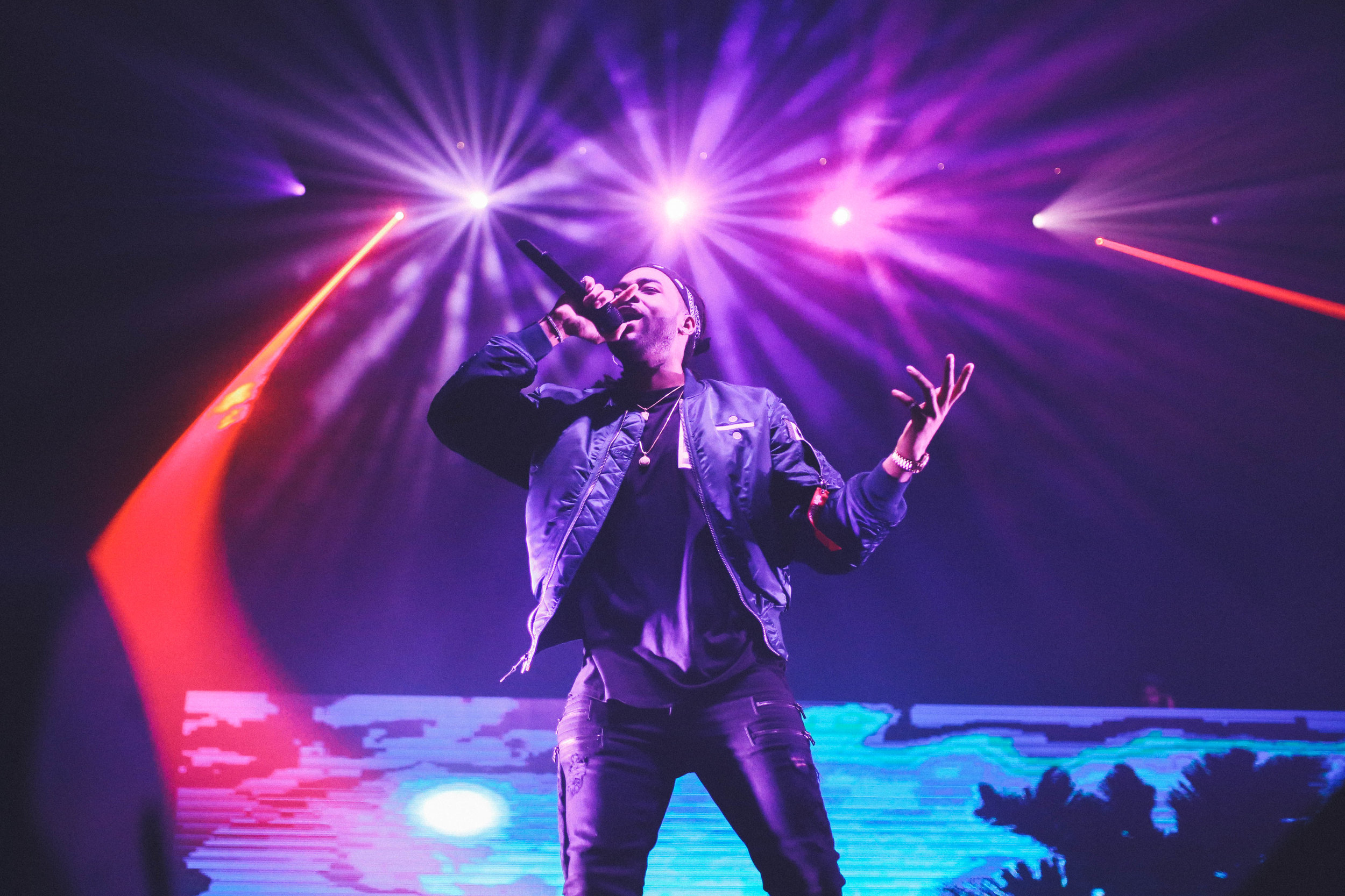 A performer on stage holding a microphone to their mouth as bright pinkish stage lights illuminate them from behind