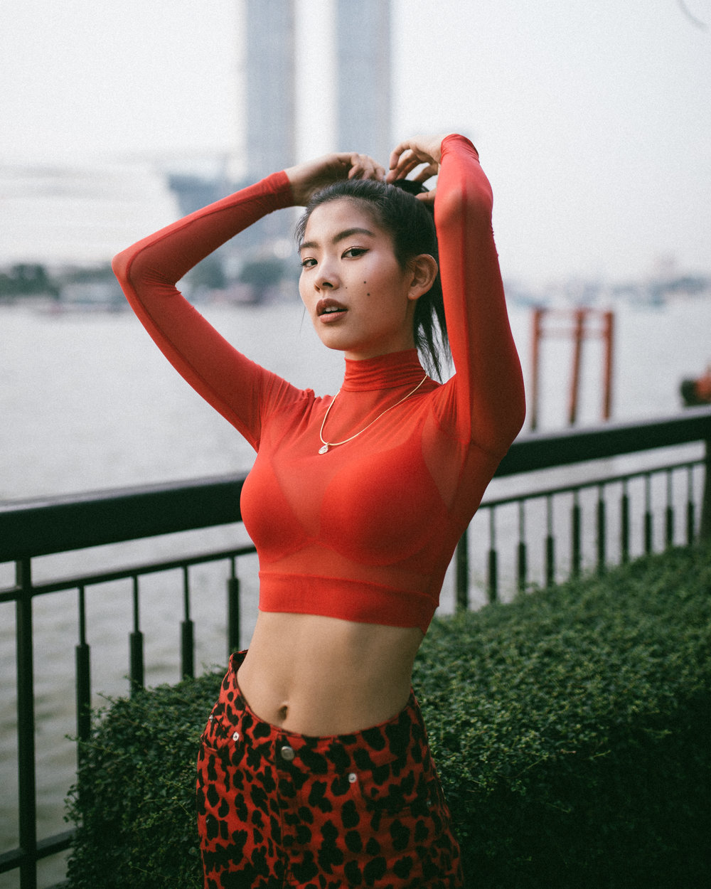 An asian woman wearing a red crop top holding both her hands up to her hair standing next to a river