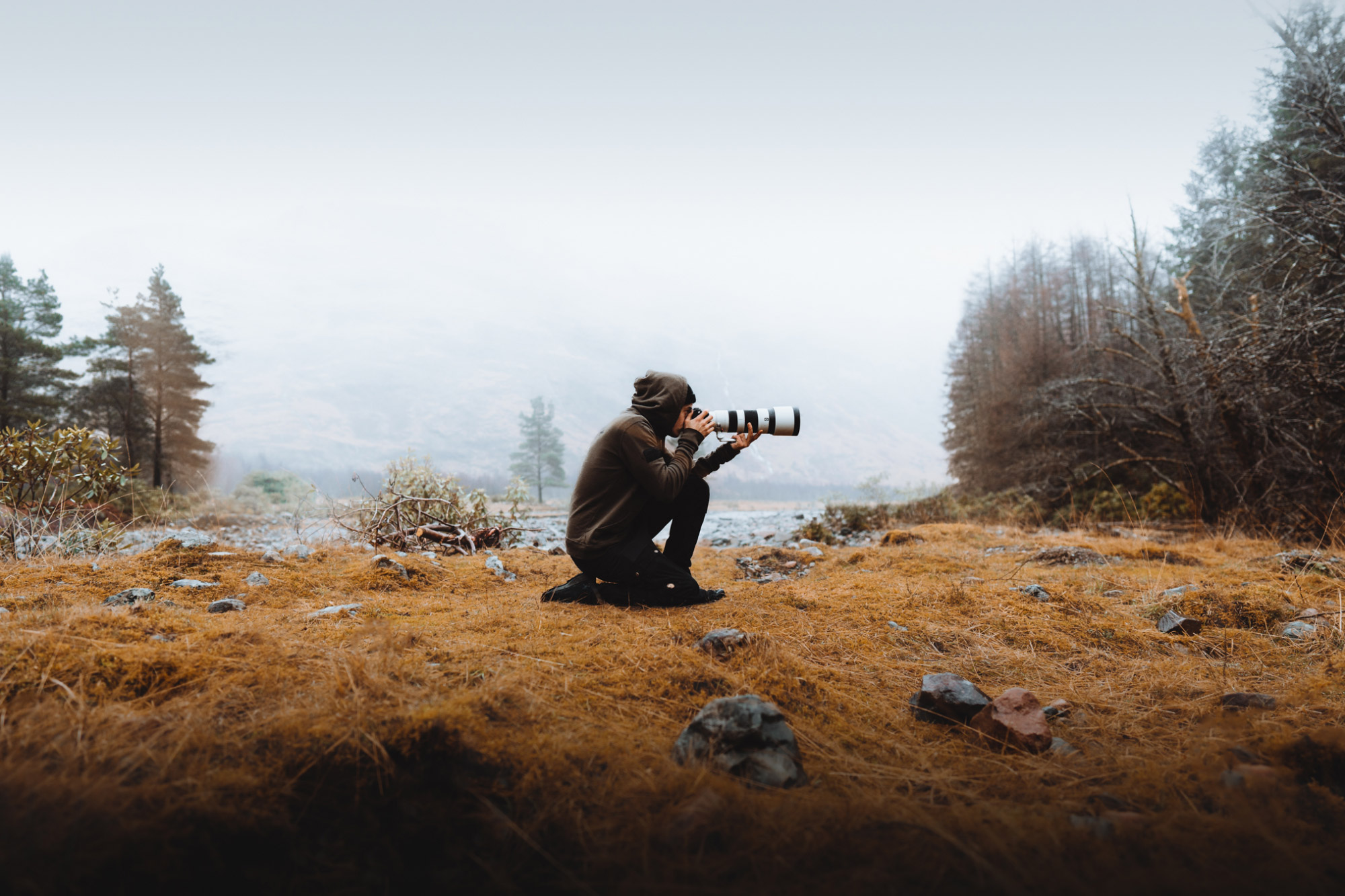 A photographer crouches whilst holding a camera in a field. There is grass in the foreground and trees and mist in the background.