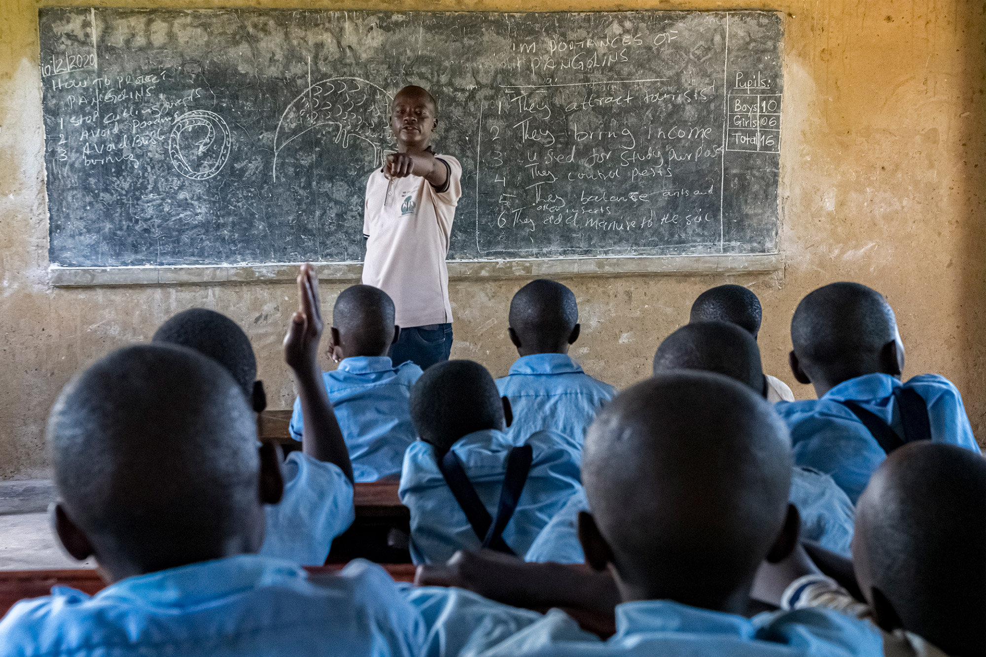 A man stands in front of a blackboard with white chalk writing on it. In front of him are several children wearing blue t-shirts.