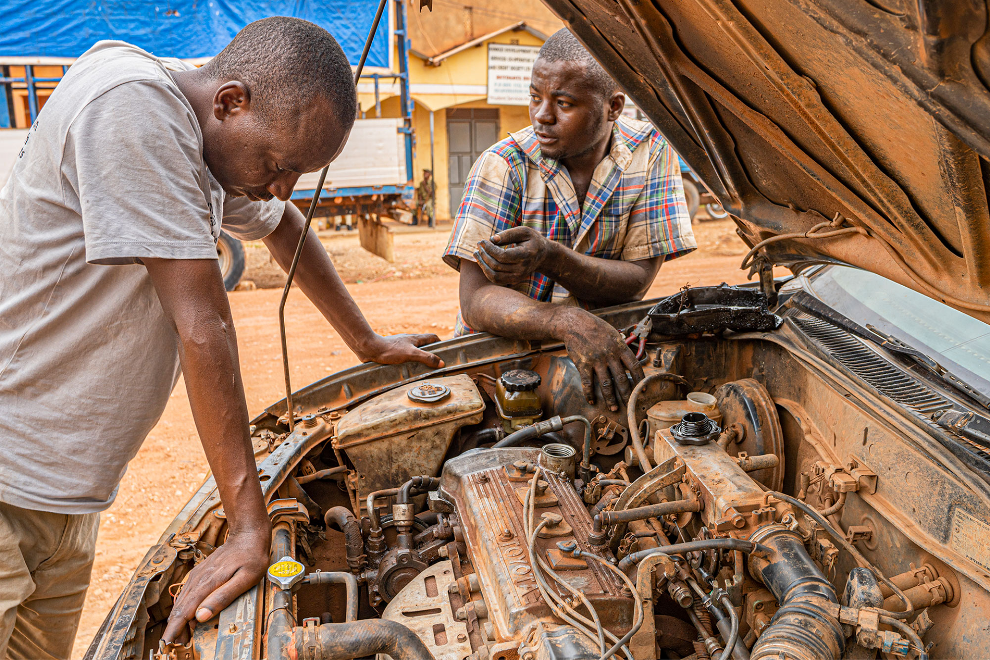 Two men stand next to car with the bonnet up. The engine inside appears to be covered in dirt.