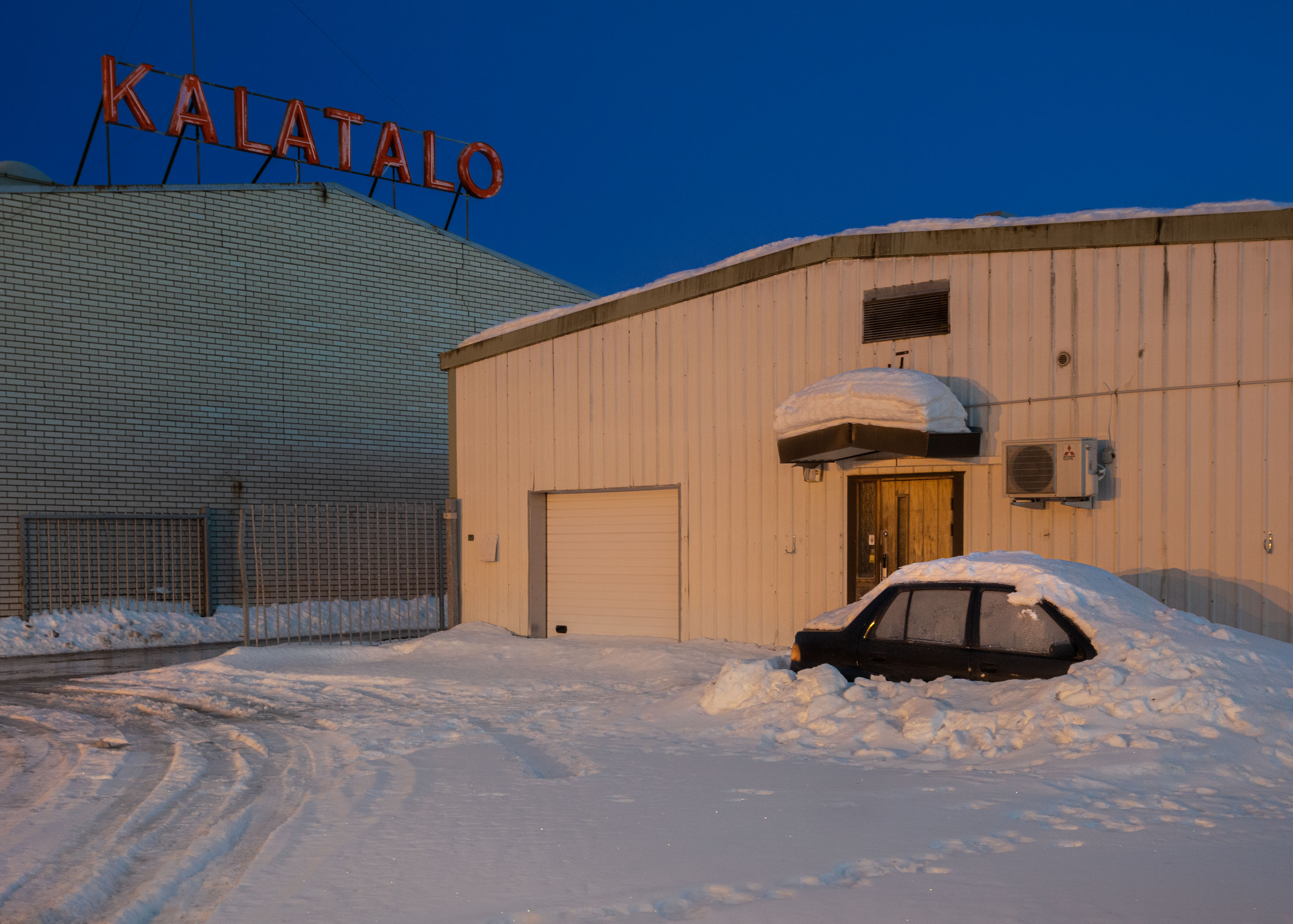 A car covered in snow near two warehouse buildings.
