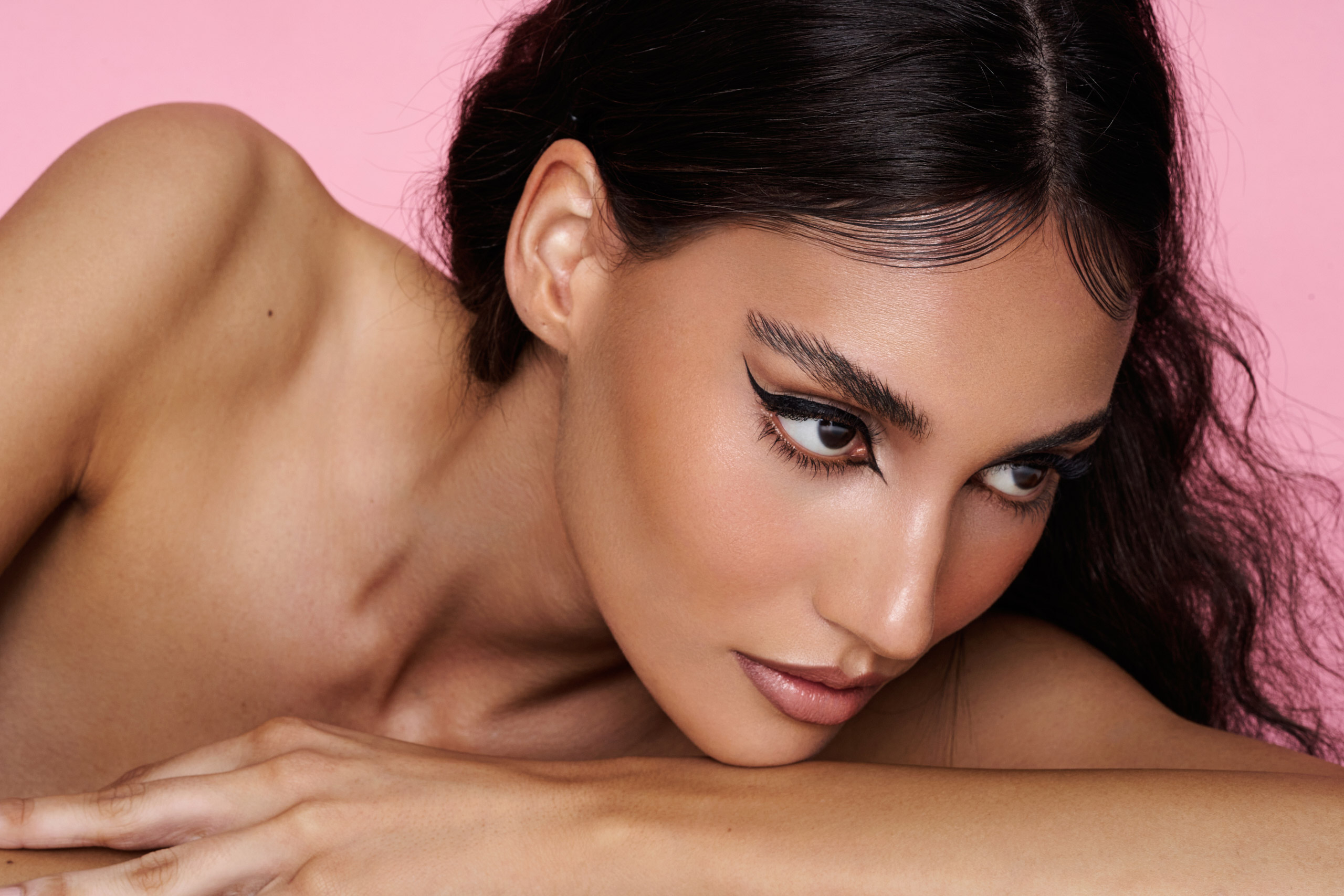 Ein Porträt des dunkelhaarigen Models Dana Mac, das sich vor einem rosafarbenen Hintergrund auf die Arme stützt, mit Make-up von Natalia Garcia