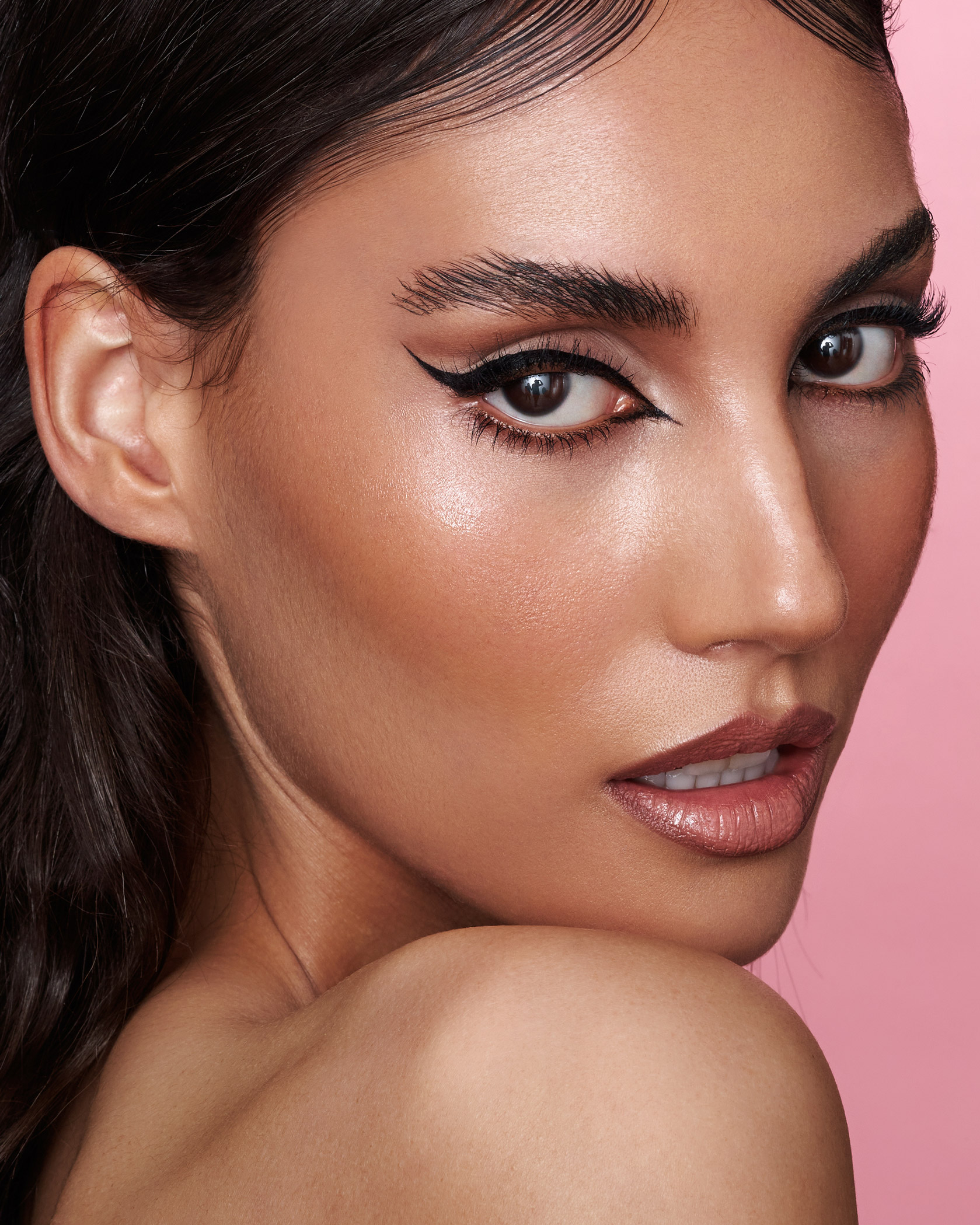 A dark-haired model, Dana Mac, looking over their shoulder against a pink background, with makeup by Natalia Garcia