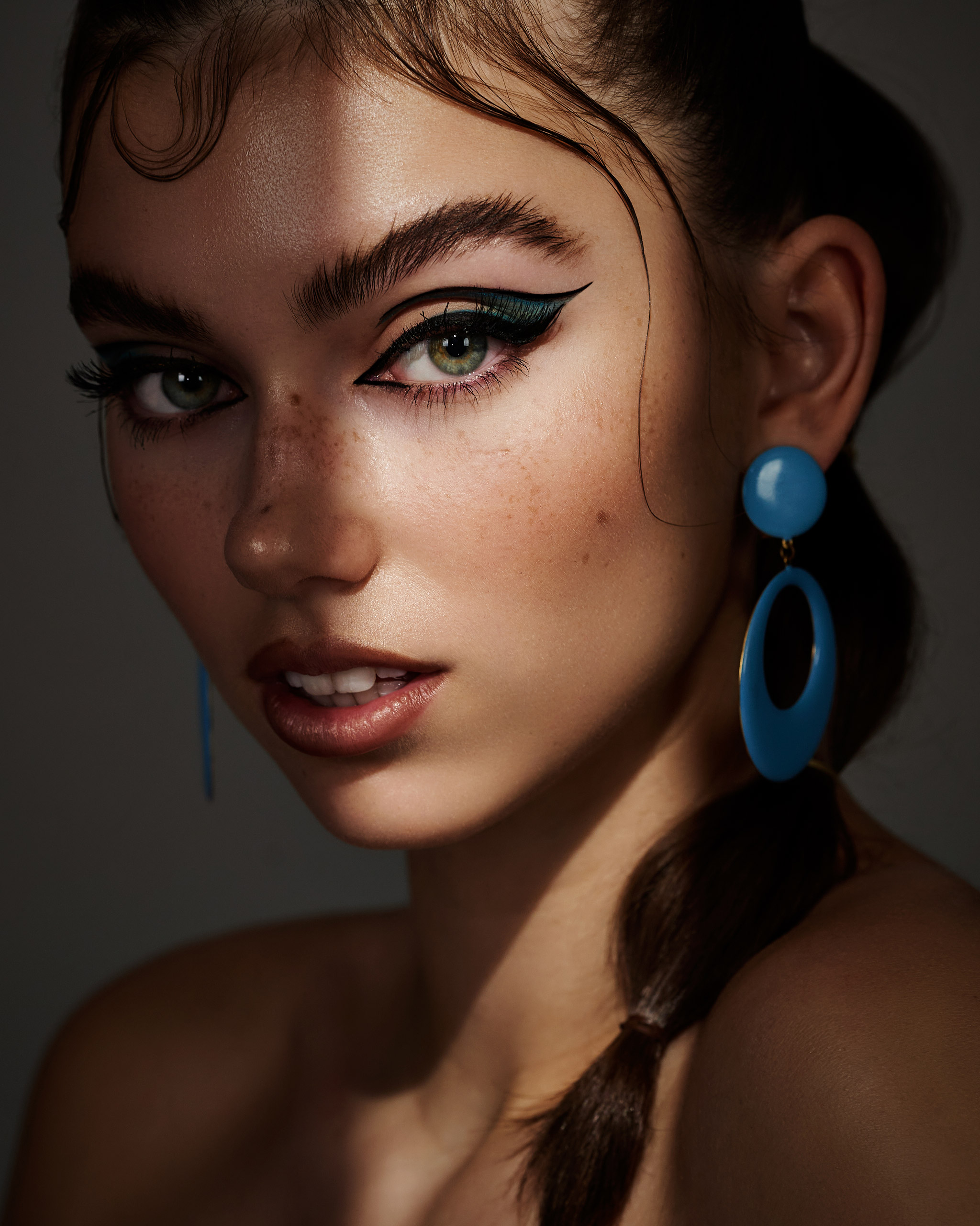 A dark-haired model, Anina, looking directly into the camera while half their face is being covered by shadow, with makeup by Natalia Garcia