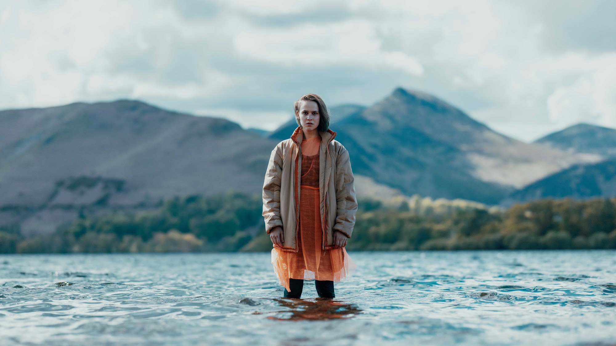 A model wearing an orange dress and a grey jacket stands in knee-level water with trees and mountains in the background.