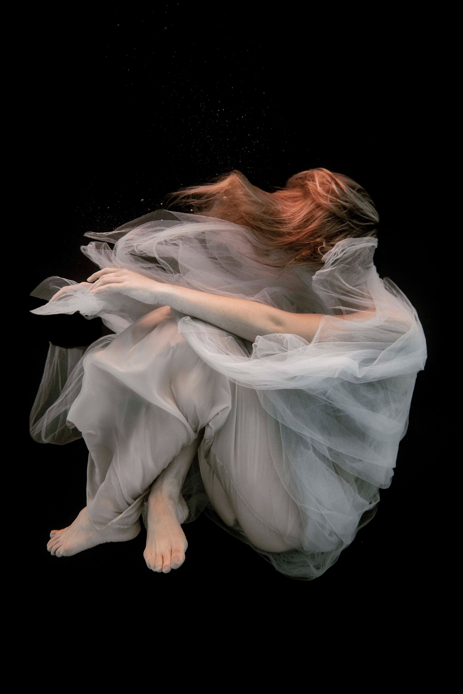 A model with red hair and a white dress underwater, the top of their dress covering their face.