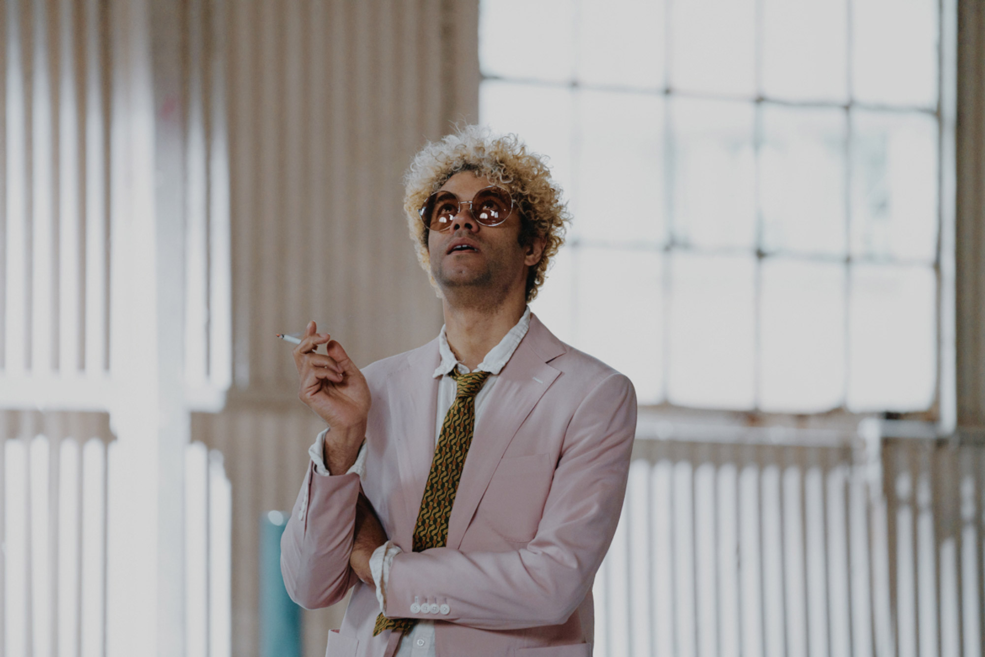 A model wearing a pink suit, blond hair and sunglasses stands holding a cigarette, with white walls and a window behind.