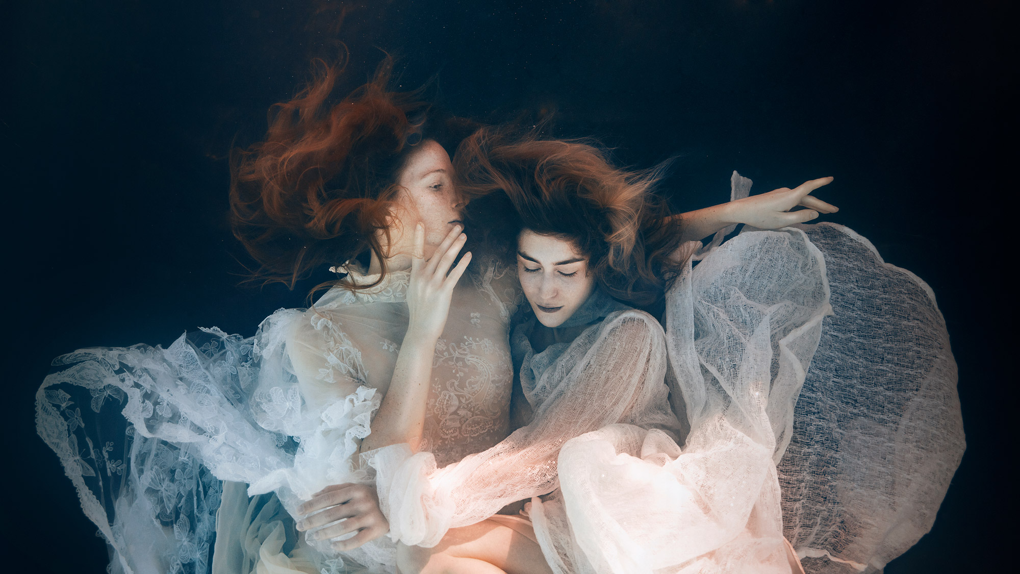 Two women with red hair and white dresses hold each other whilst under water.