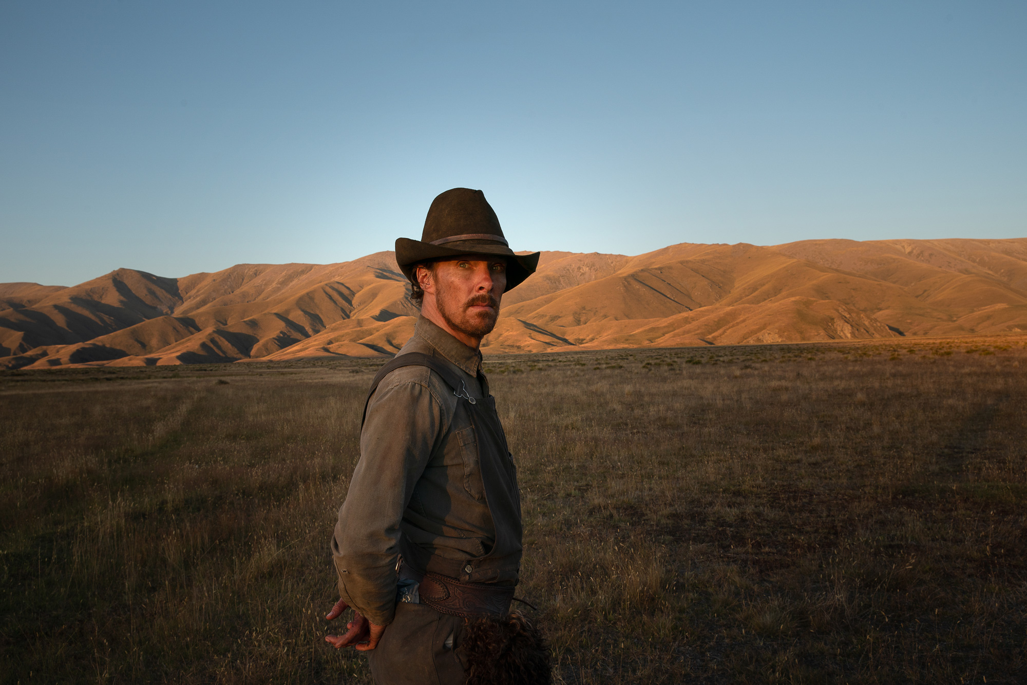 Benedict Cumberbatch dressed as Phil Burbank stands with his hands behind his back on the set of the film The Power of the Dog there are mountains and fields in the background.