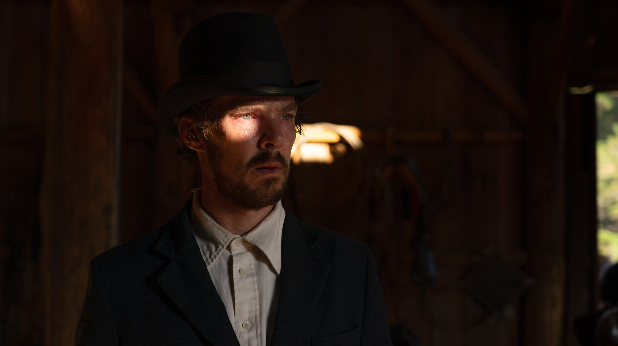 Benedict Cumberbatch dressed as Phil Burbank stands with his hands behind his back on the set of the film The Power of the Dog there are mountains and fields in the background.