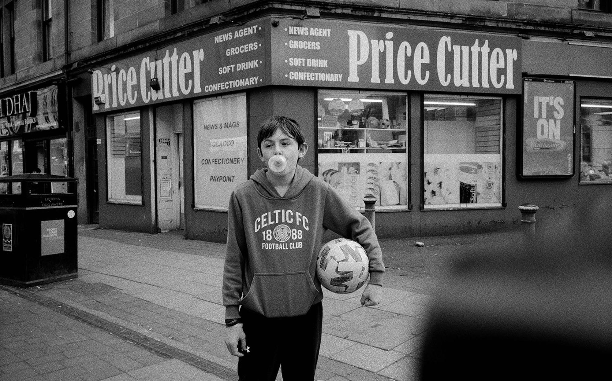 
Un jeune se tenant à l'extérieur de Price Cutter avec un ballon de football, faisant une bulle de chewing-gum.

