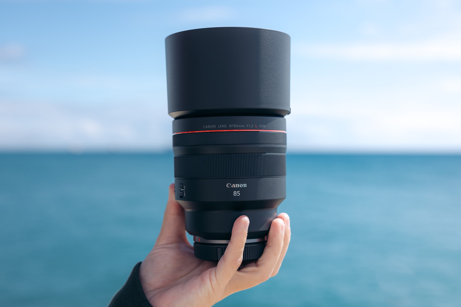 A hand holds a Canon RF 85mm f/1.2 L USM lens, with blue sky and sea in the background.