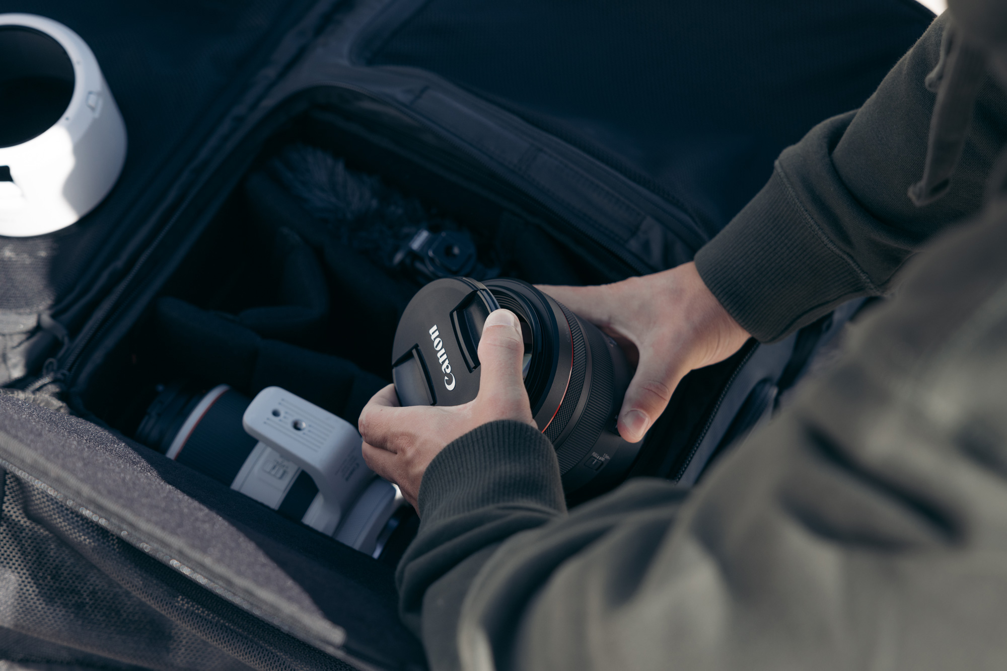 A photographer wearing a black jumper reaches into a camera back to pick a Canon RF 85mm f/1.2 L USM lens.