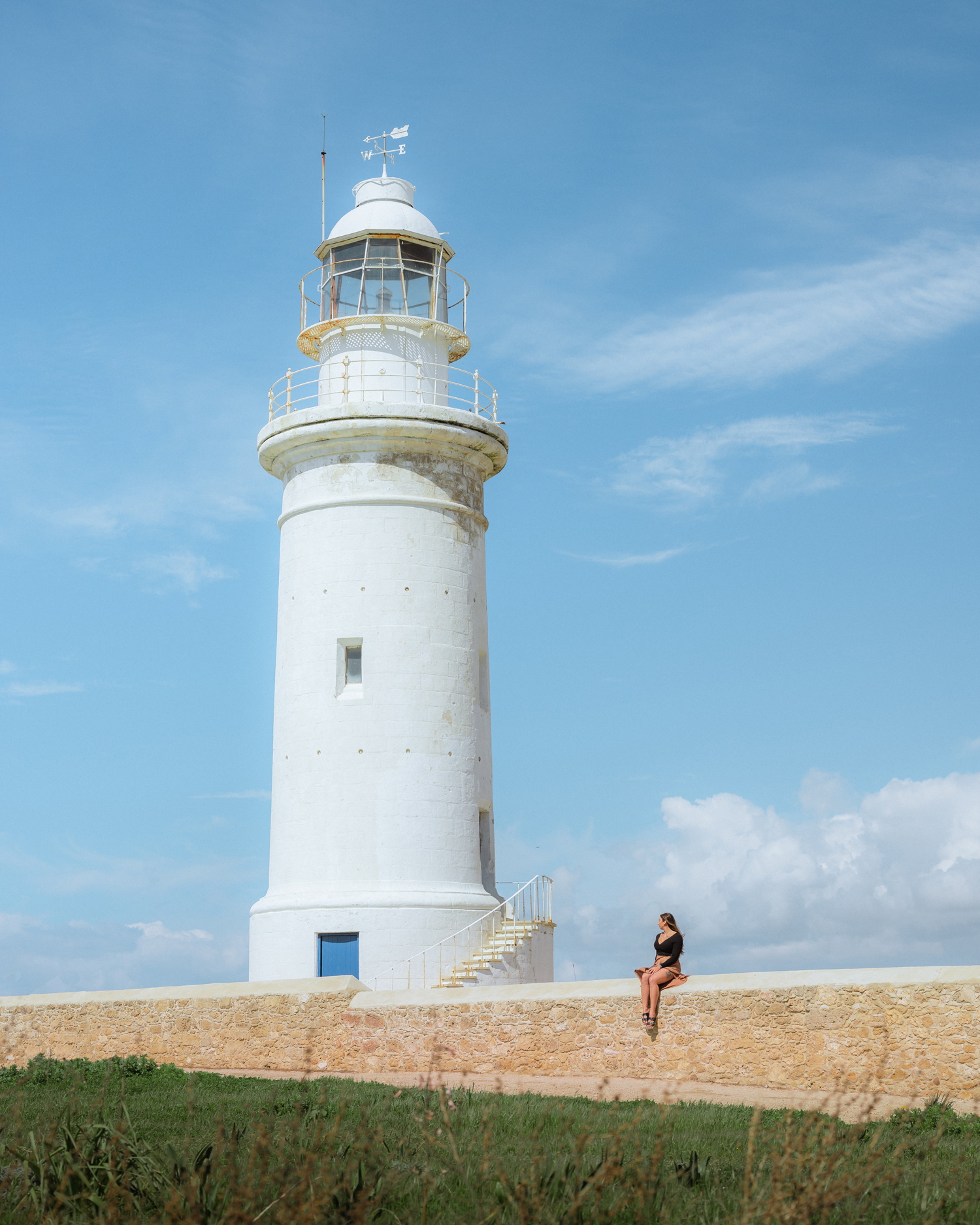 A person sits on a wall near a white lighthouse, with grass in the foreground and blue sky in background.