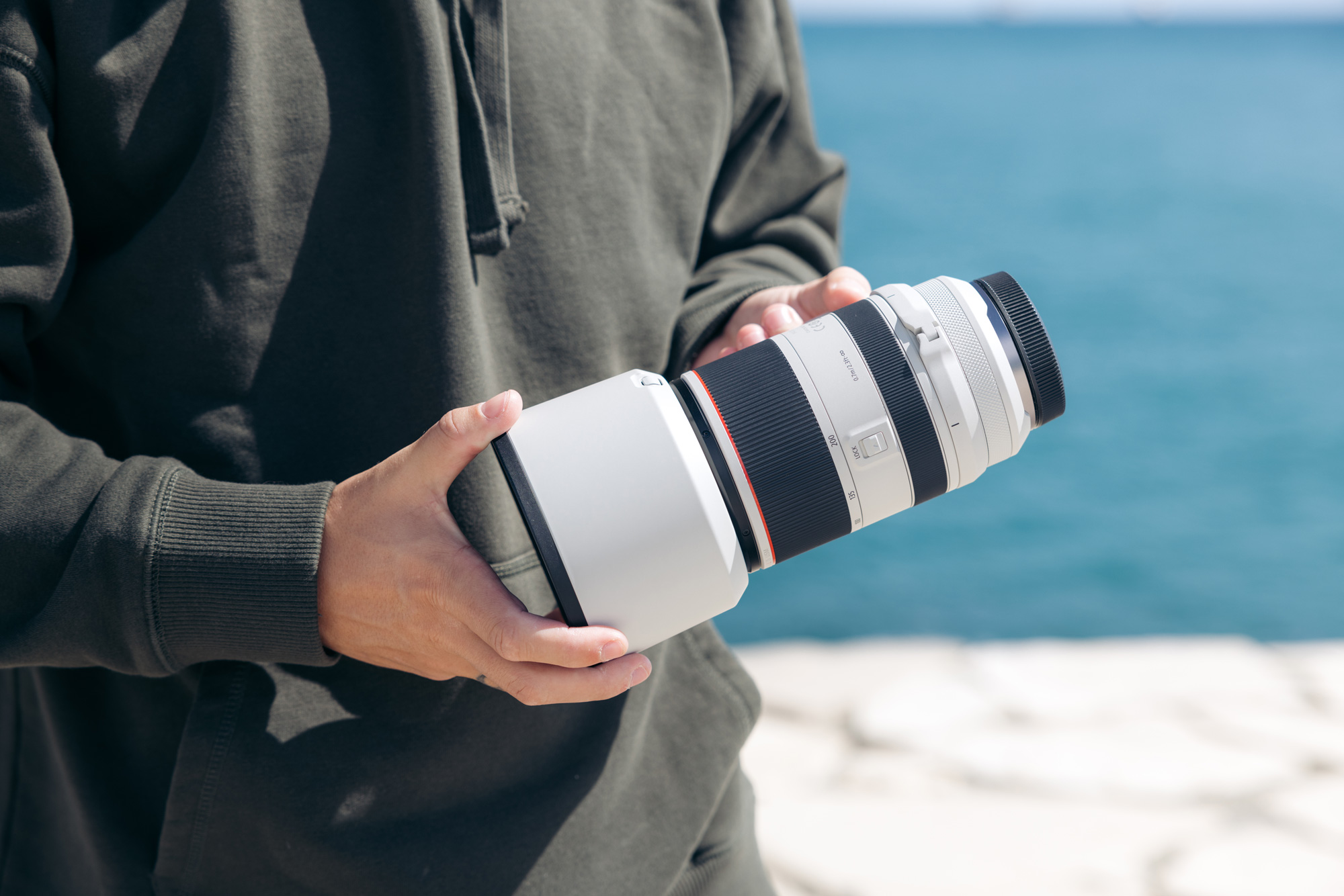 A photographer wearing a black hoodie hold the Canon RF 70-200mm f/2.8 lens with water in the background.