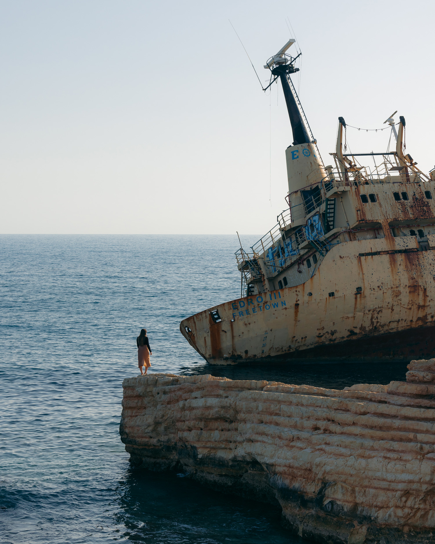 A person stands on a wall behind a ship wreck with blue sky and sea behind them.