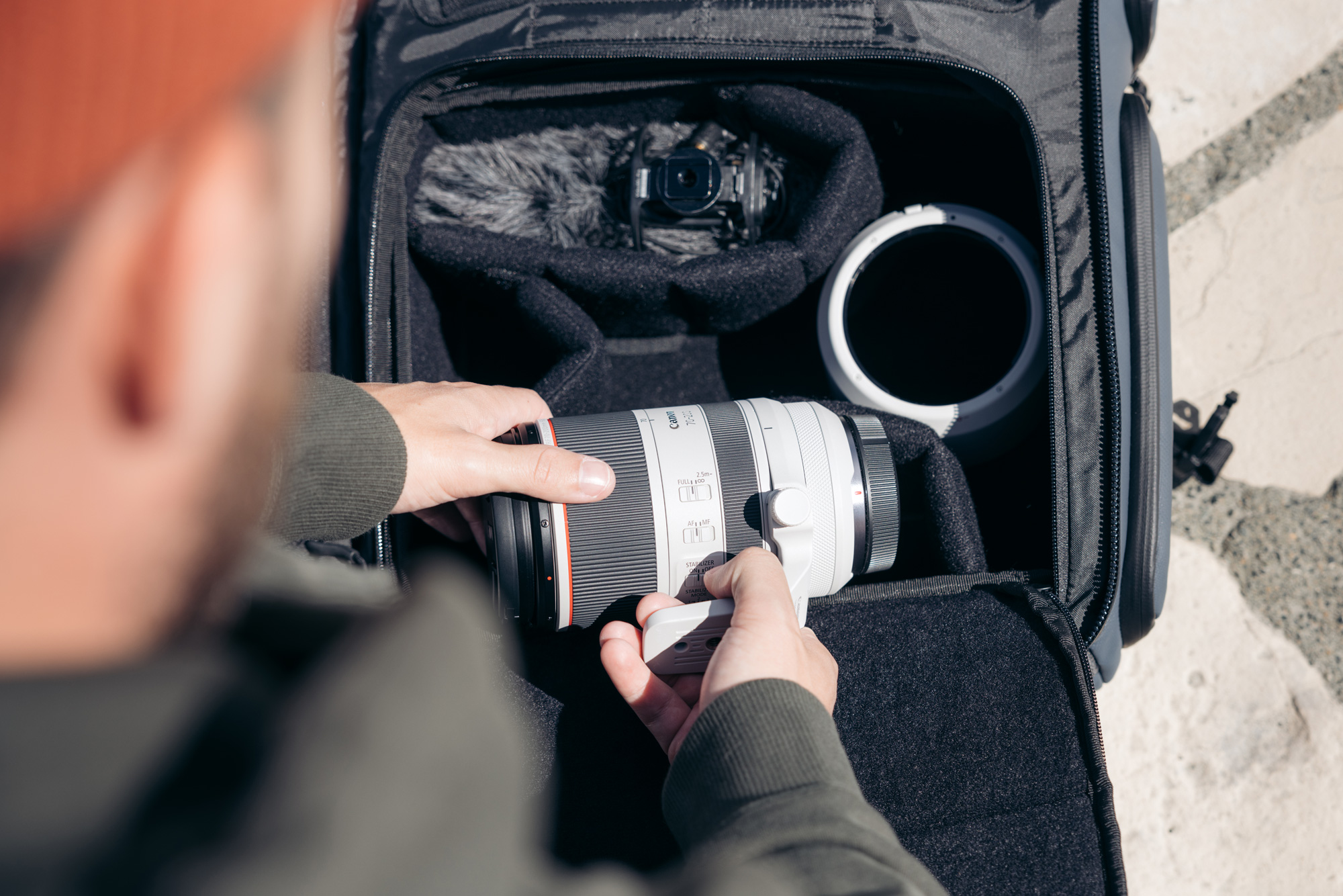 A photographer wearing a black jumper reaches into a camera back to pick up a Canon RF 70-200mm f/2.8 lens.