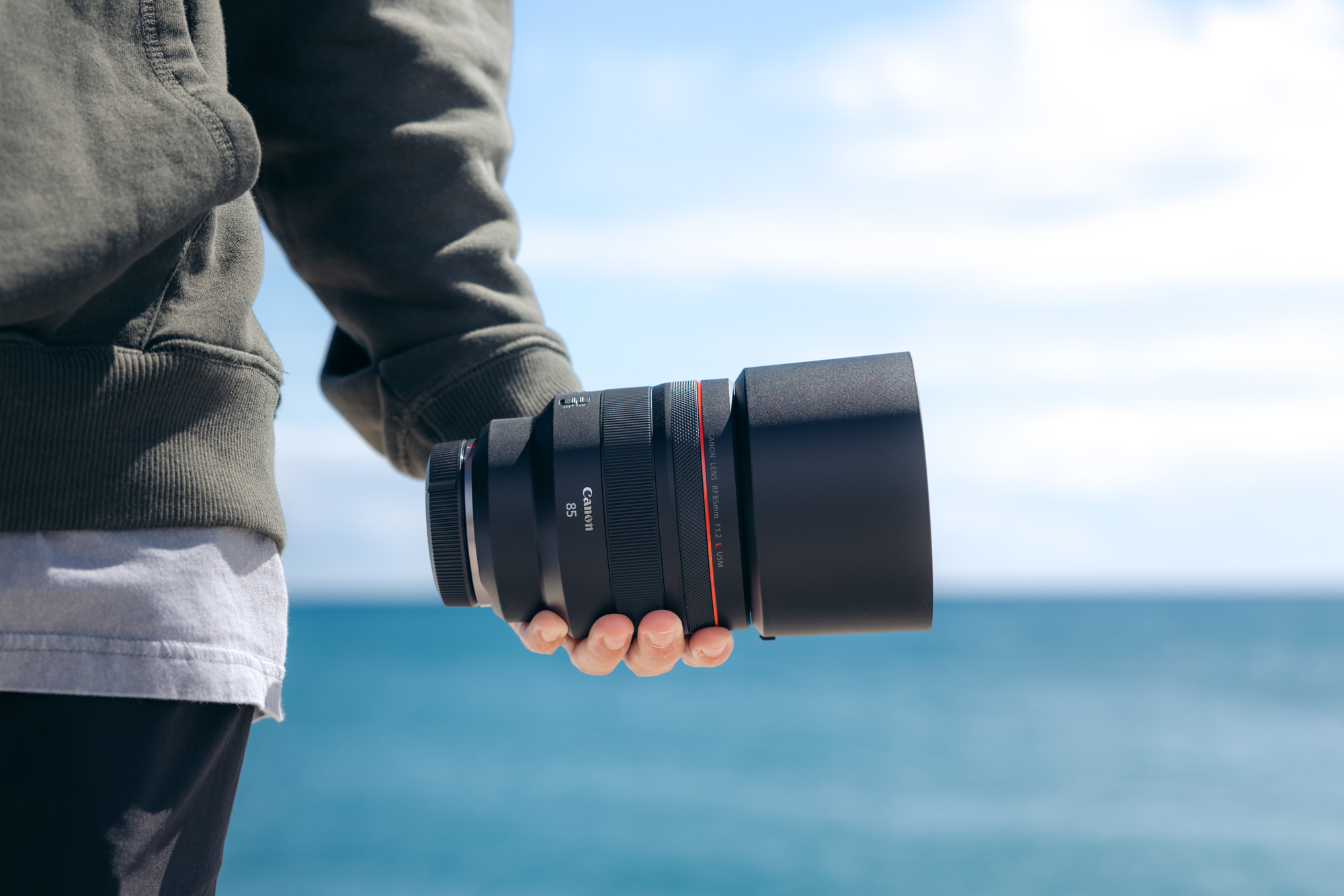 A hand holds a Canon RF 85mm f/1.2 L USM lens, with blue sky and sea in the background.