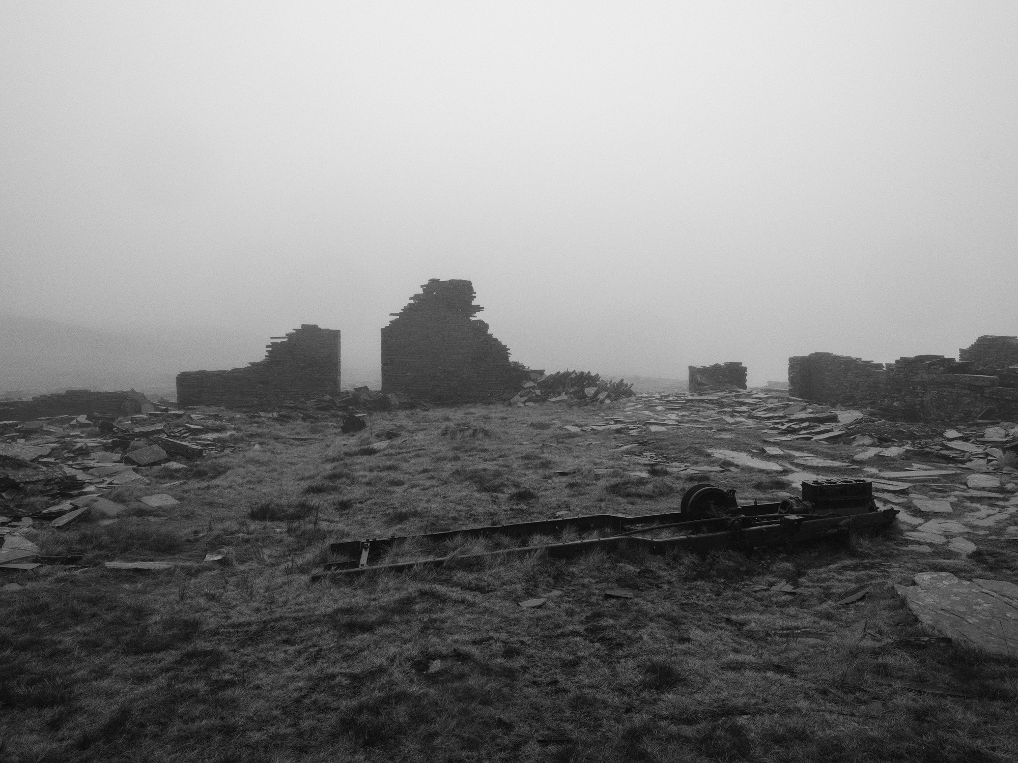 A black and white image of an abandoned mine. An old piece of machinery sits on a grassy area in the foreground, in the background, there are a pile of slate tiles.