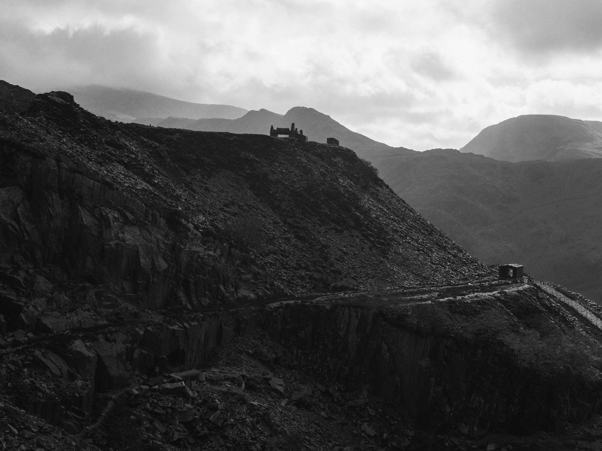 A black and white image of a hillside. There is a pathway halfway up the hill with a cabin at the end. At the top, there is a cabin half hidden. There are mountains in the background