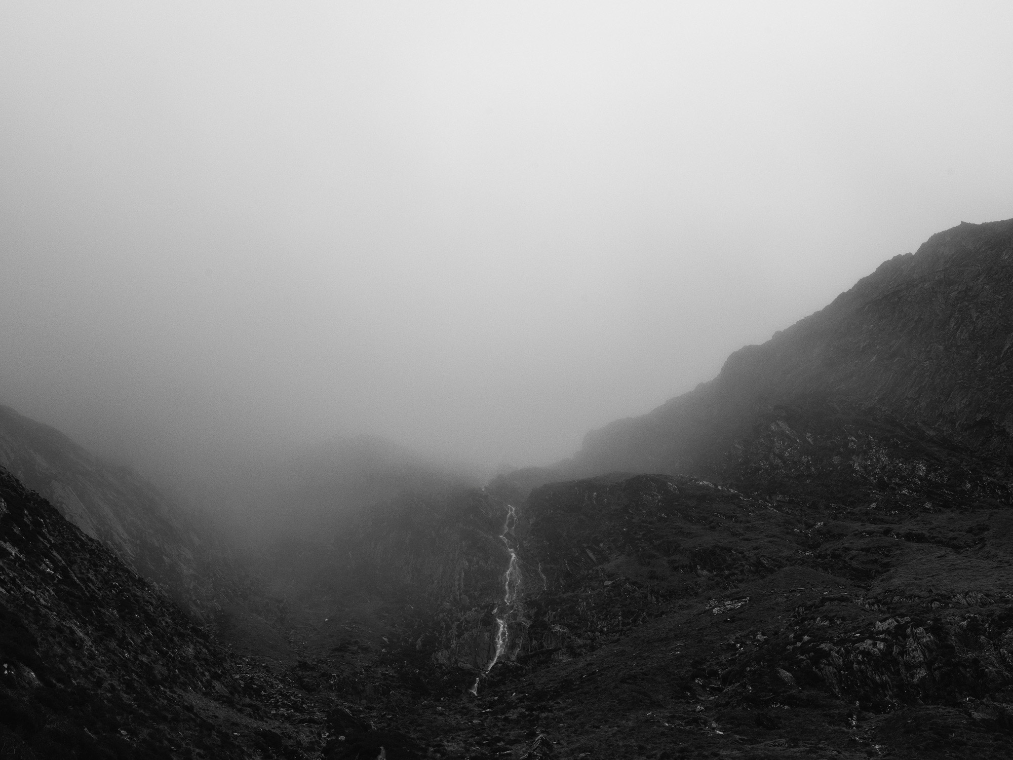 A block and white image of a river running through a valley with mountains either side. The background is hidden by clouds and fog.