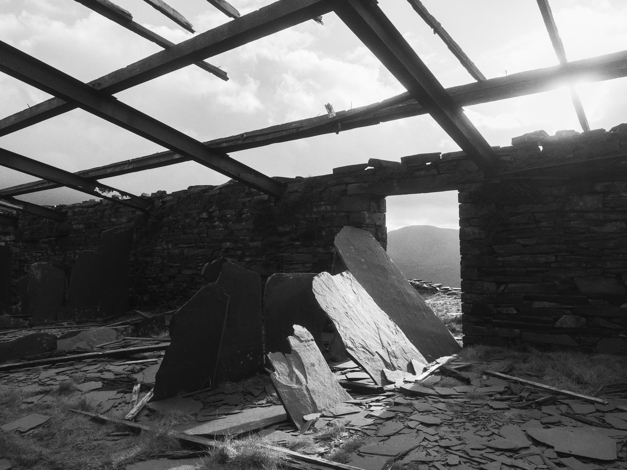 A black and white image of a brick wall with a door missing. In the foreground there are pieces of flint, some of which are standing up. Above the wall there are wooden beams and in the background a mountain.
