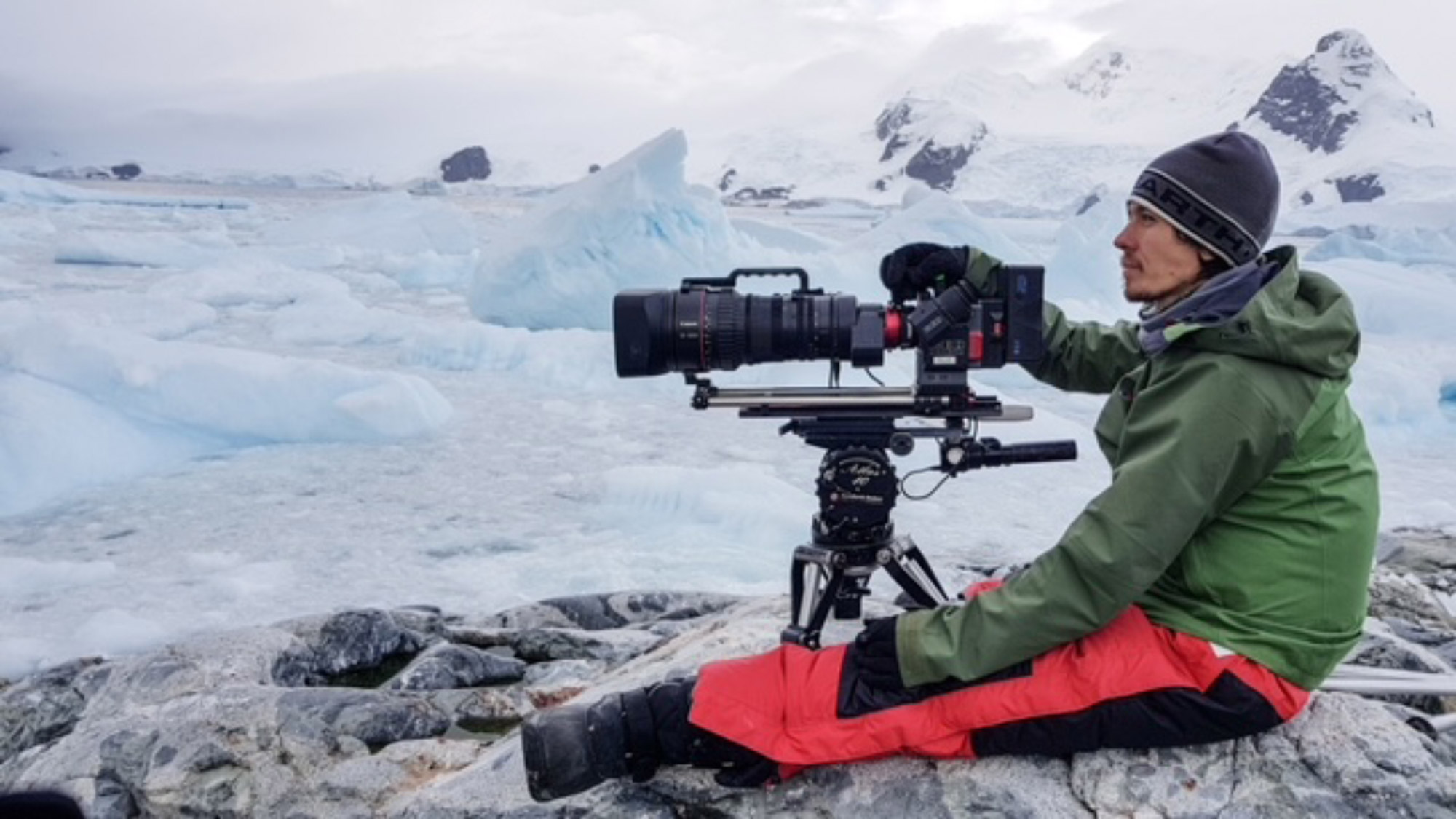 Alex Vail sitzt mit der Kamera in der Hand auf dem Boden, mit Eisbergen im Hintergrund